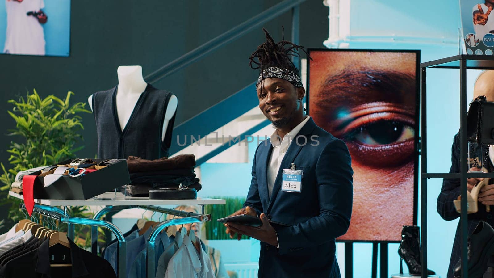 African american worker managing clothes stock on hangers, working on inventory using tablet in shopping center. Male store assistant in a suit preparing fashion collection on display. Camera A.