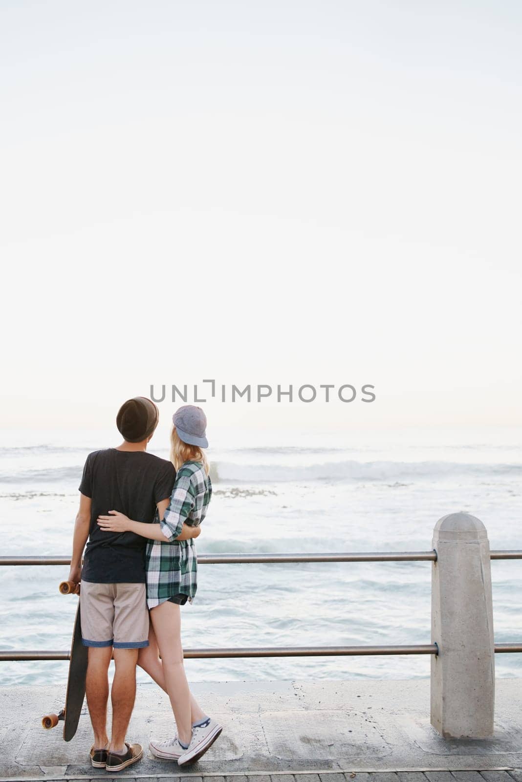 Couple, rear view and hug on beach for travel with mockup space, adventure and promenade date. Gen z skater, people and skateboard on boardwalk for bonding, embrace and outdoor fun with morning waves by YuriArcurs