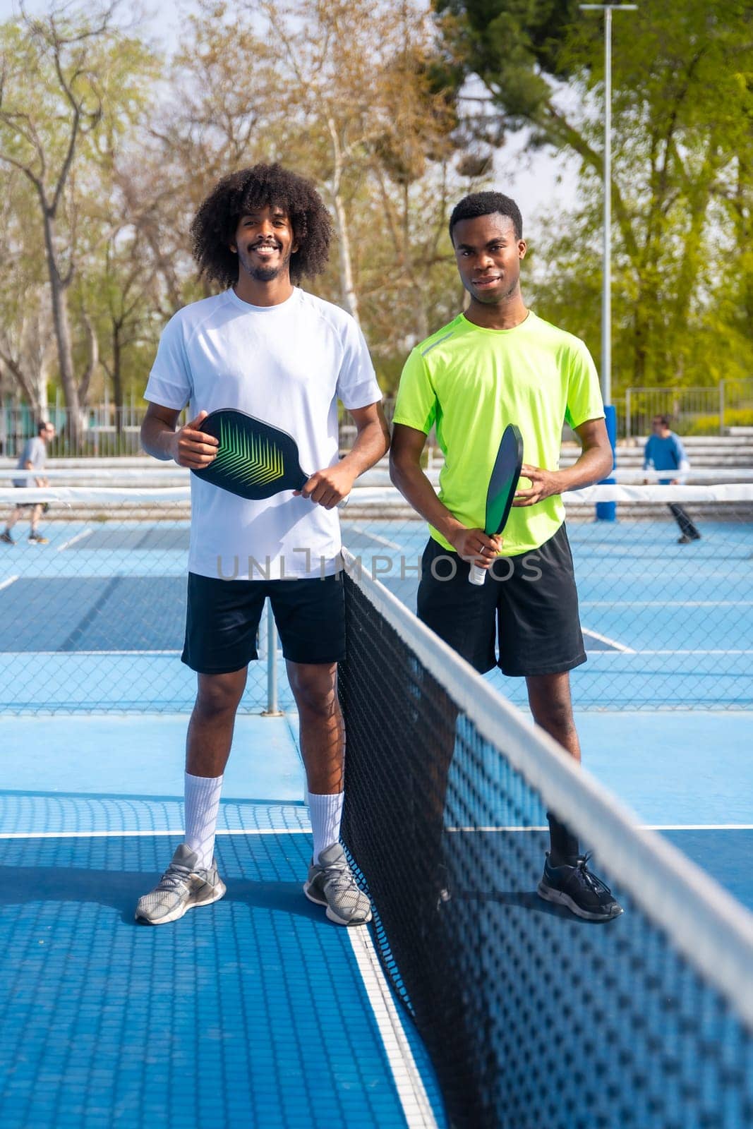 Vertical Portrait of two young african american pickleball players standing looking at camera holding pickleball equipment