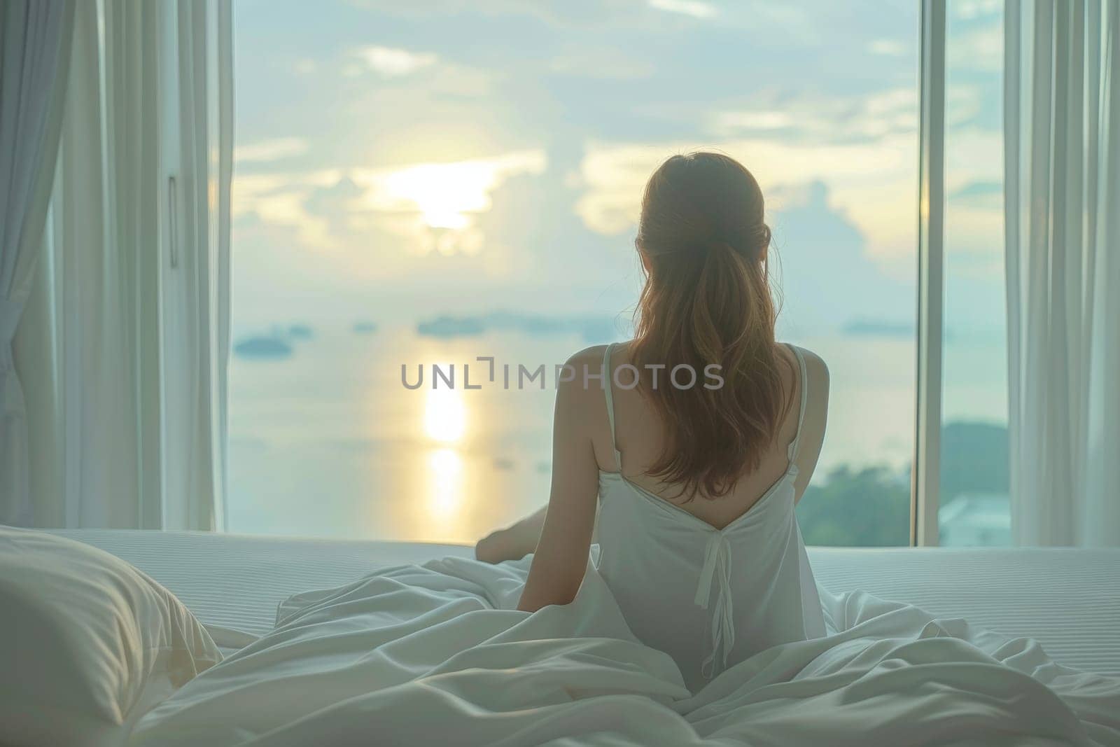 A woman is sitting on a bed with her back to the camera. She is wearing a white robe and she is relaxed. The room has large windows that let in natural light