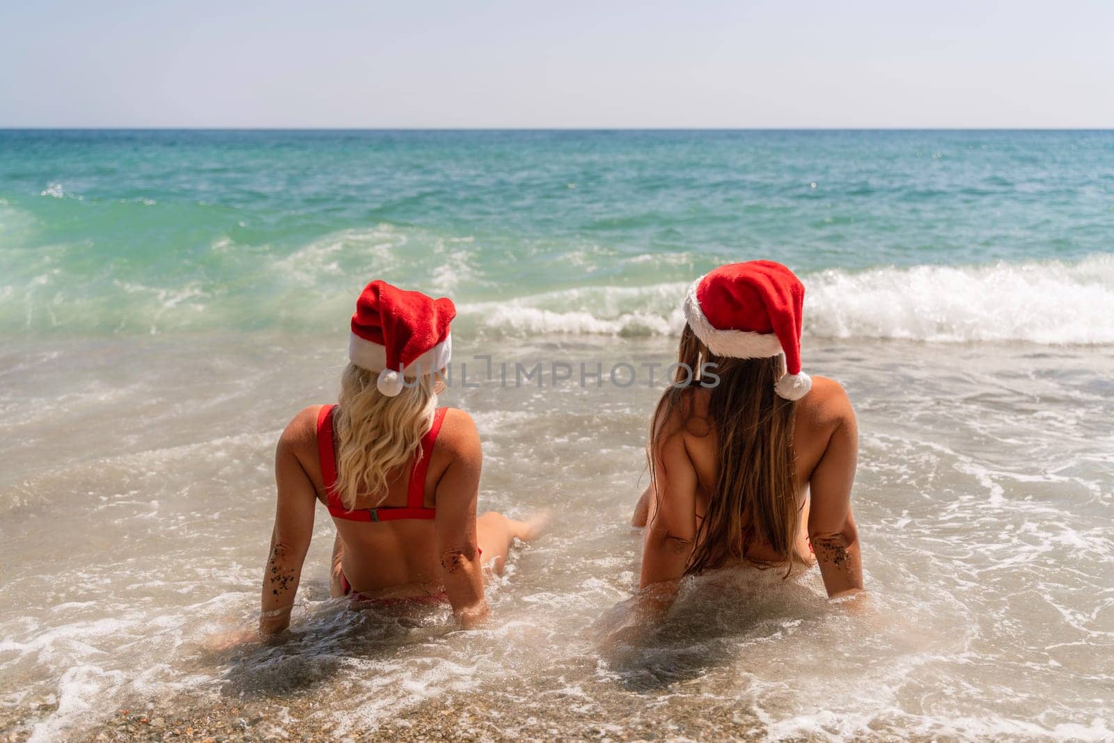 Women Santa hats ocean play. Seaside, beach daytime, enjoying beach fun. Two women in red swimsuits and Santa hats are enjoying themselves in the ocean waves. by Matiunina