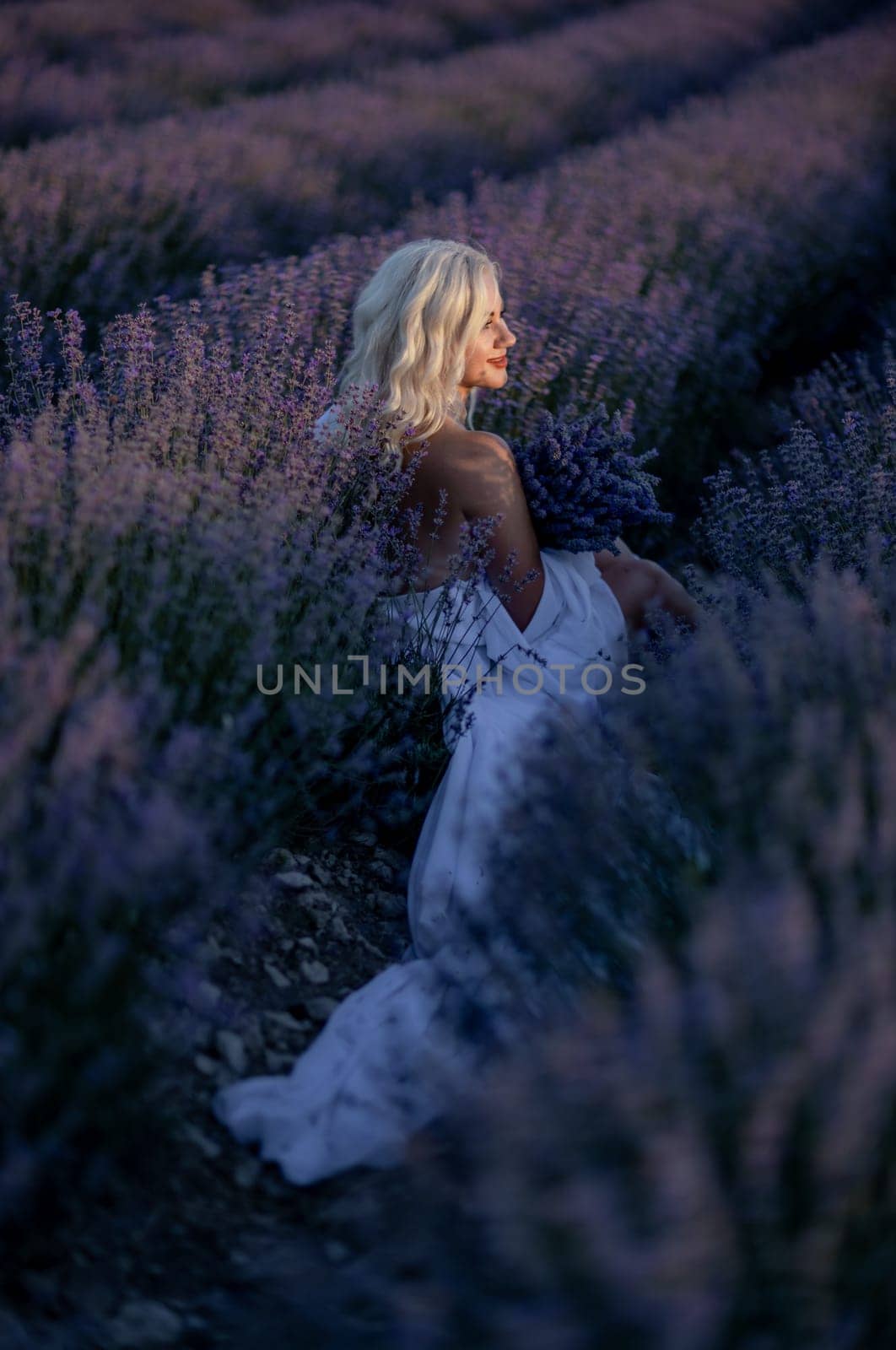 Blonde woman poses in lavender field at sunset. Happy woman in white dress holds lavender bouquet. Aromatherapy concept, lavender oil, photo session in lavender by Matiunina