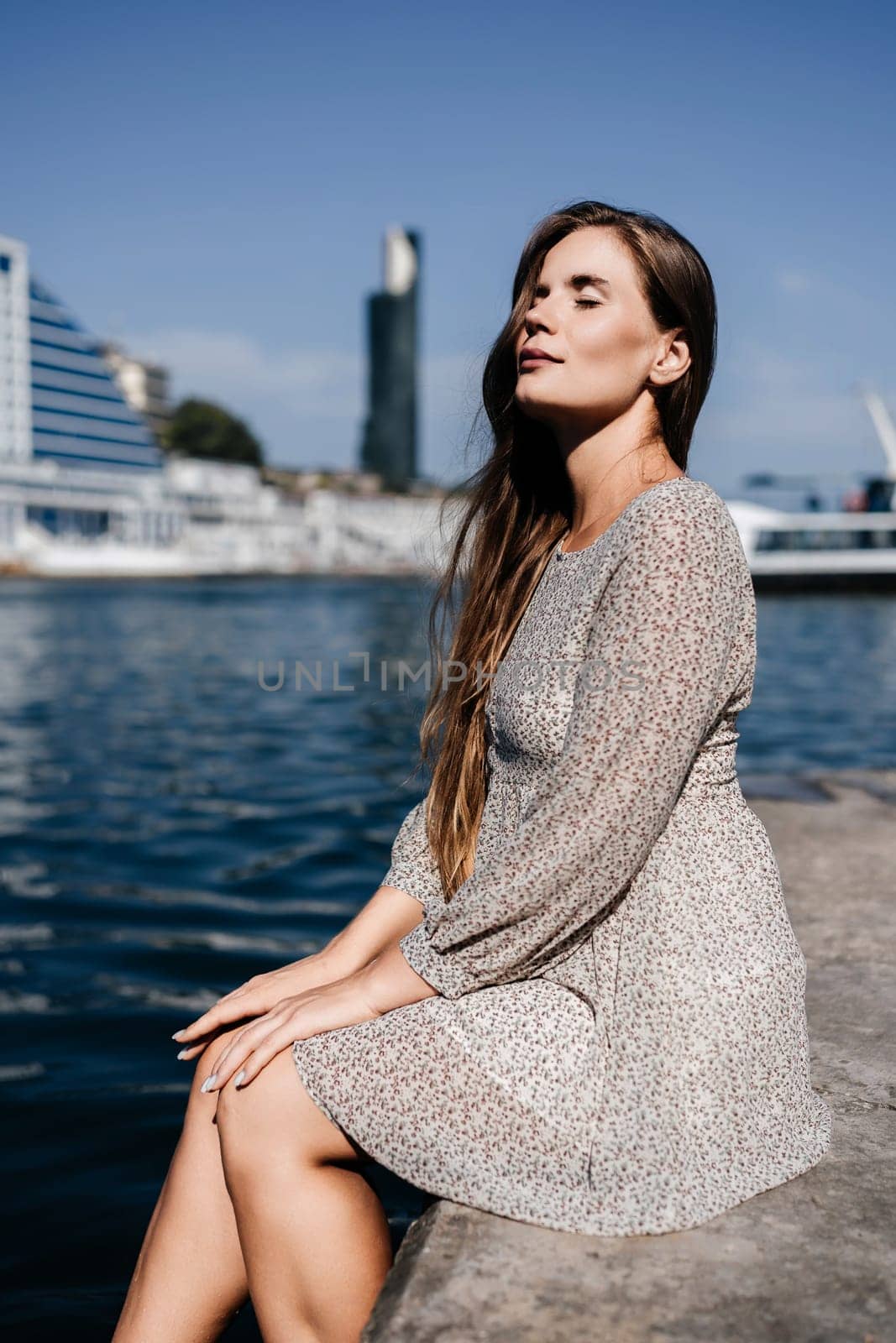 A woman with long hair is sitting on a dock by the water. She is wearing a white dress and she is looking at the water. The scene has a calm and peaceful mood