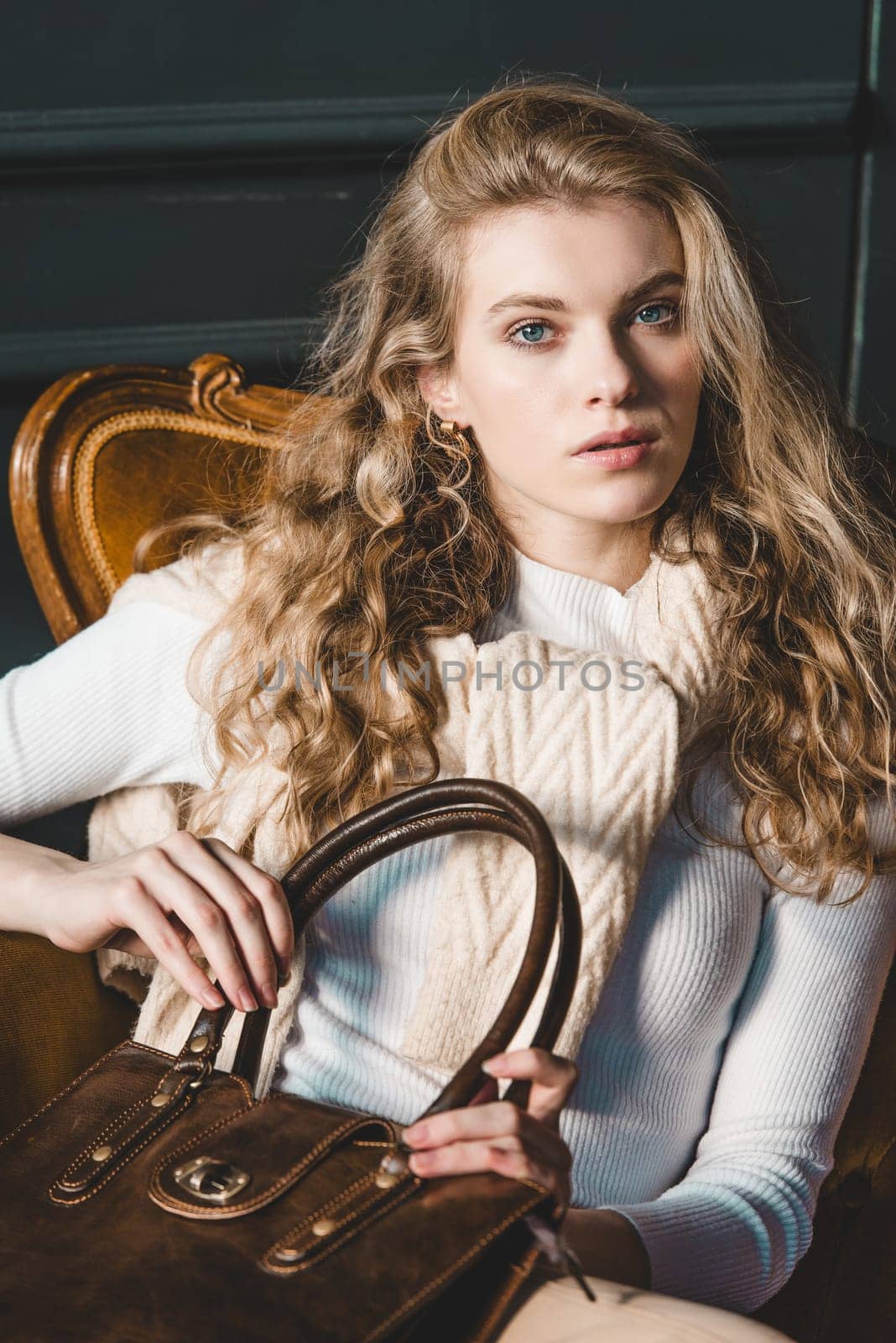 beautiful curly blond hair woman posing with a small shopper brown bag in a vintage chair. Model wearing stylish white sweater and classic trousers