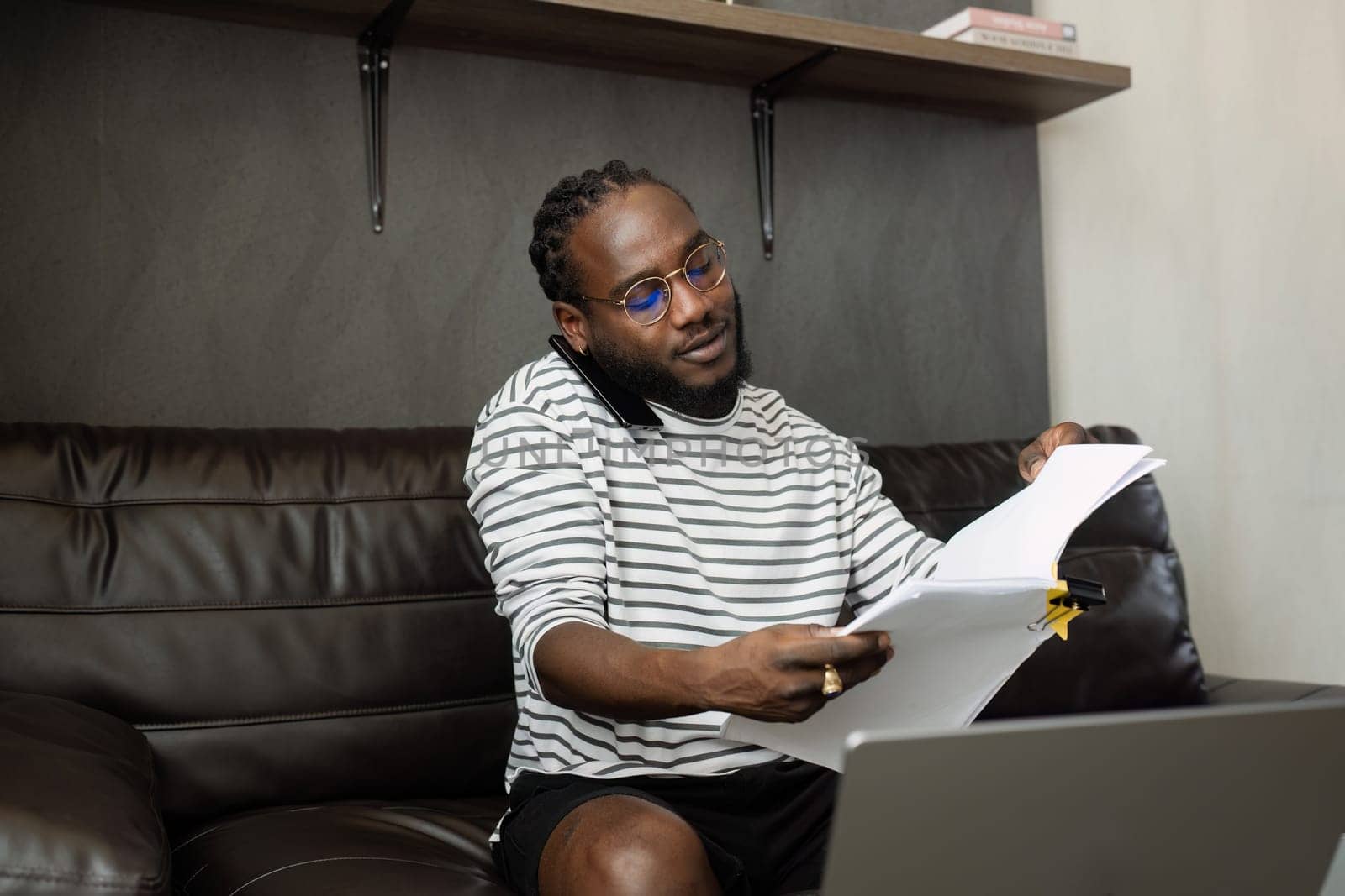 African American man working with document and laptop while talking on mobile at home on couch. work from home concept.