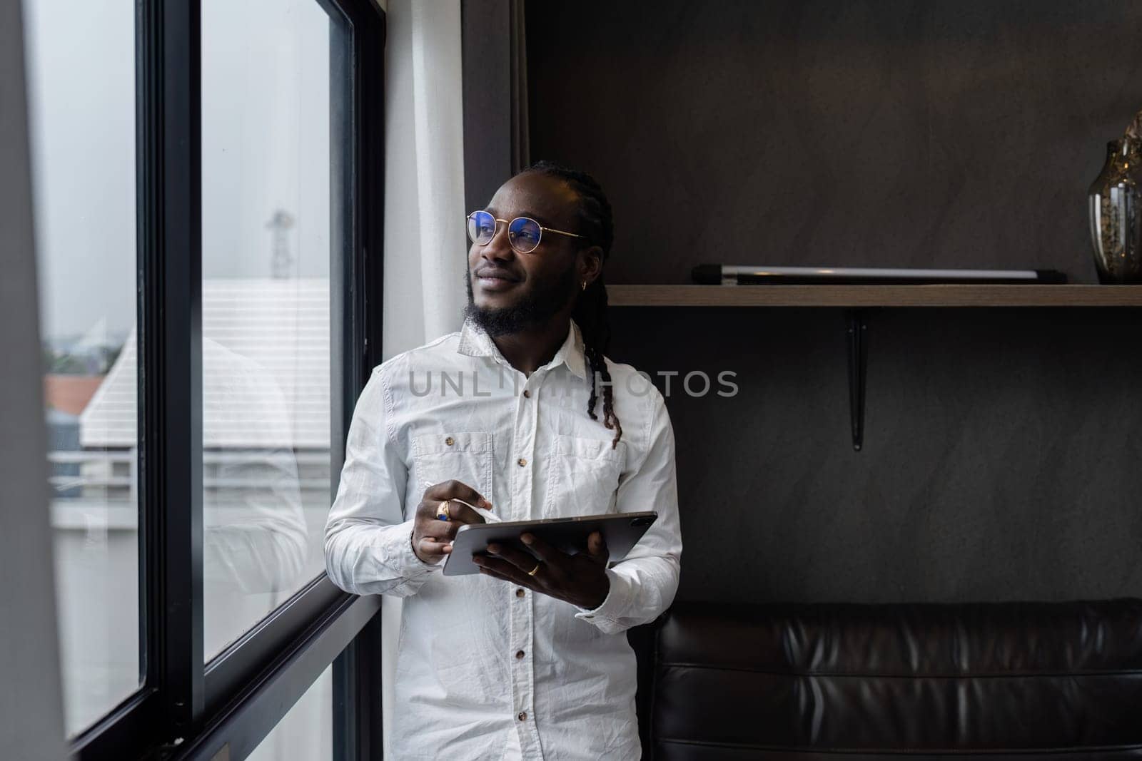 African American business man working at home on weekend, holding tablet and standing looking out the window.