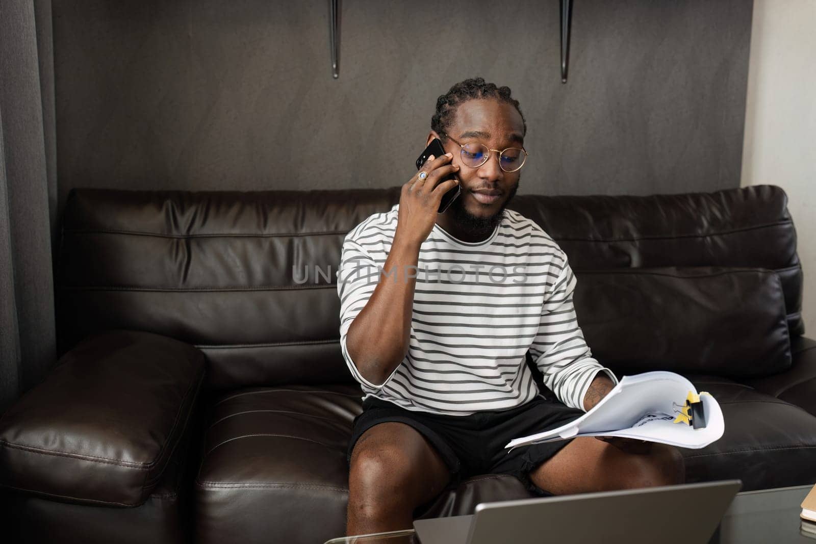 African American man working with document and laptop while talking on mobile at home on couch. work from home concept.