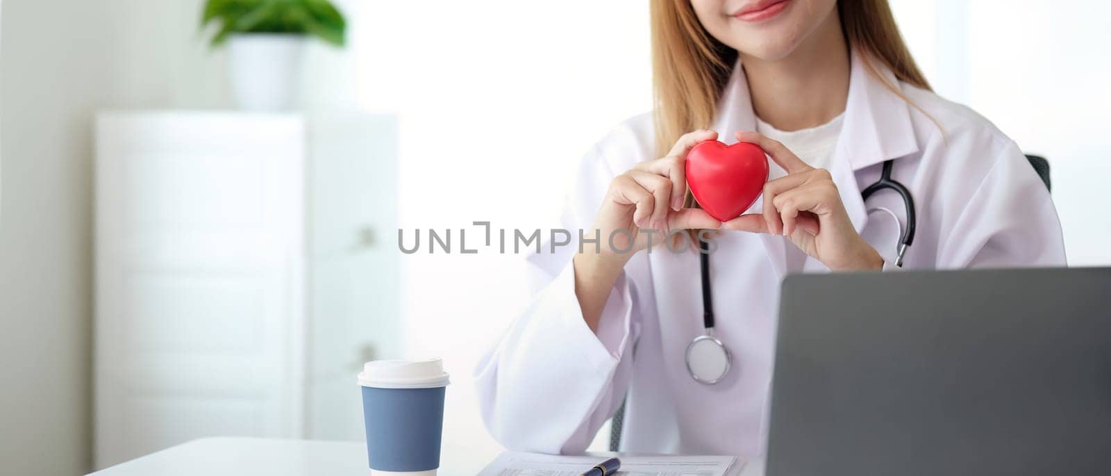 Happy young doctor woman holding red heart shape object, looking at camera with smile. Positive practitioner, cardiologist.