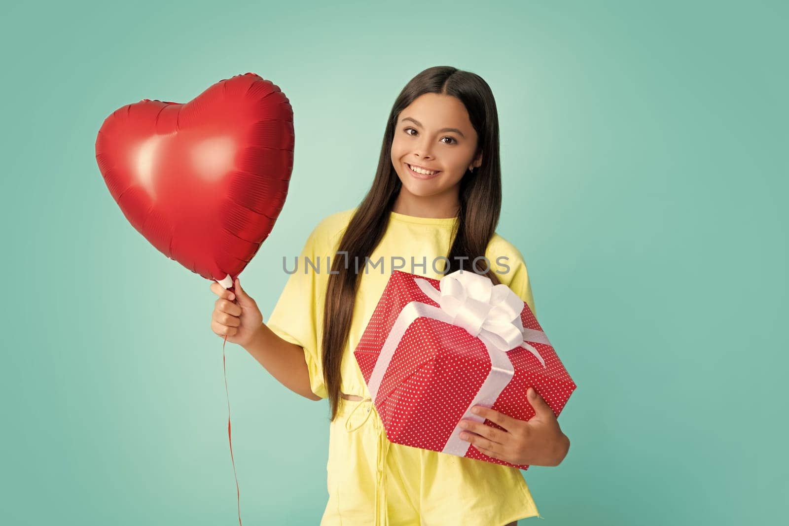 Teenager child holding gift box on blue isolated background. Gift for kids birthday. Christmas or New Year present box. Happy girl face, positive and smiling emotions. Love valentines, heart balloon