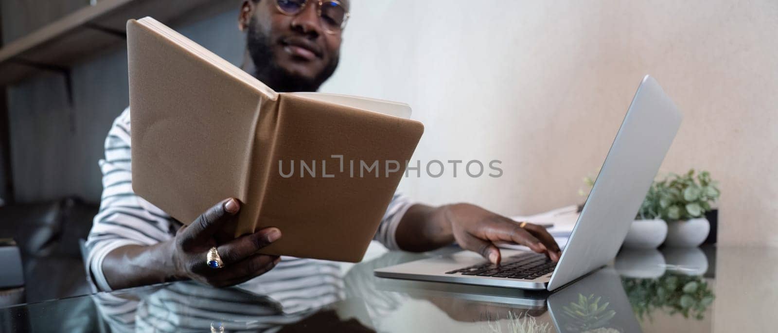 Young african man using laptop at home, black male looking at read book relaxing on leisure with work sit on glass table in living room by nateemee