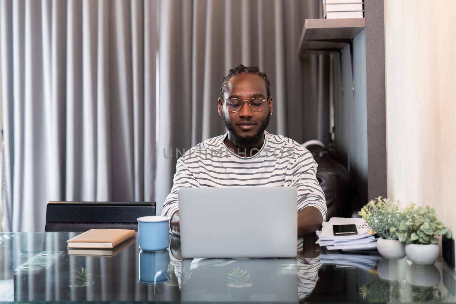 African American man working with laptop computer remote while sitting at glass table in living room. Black guy do freelance work at home office by nateemee