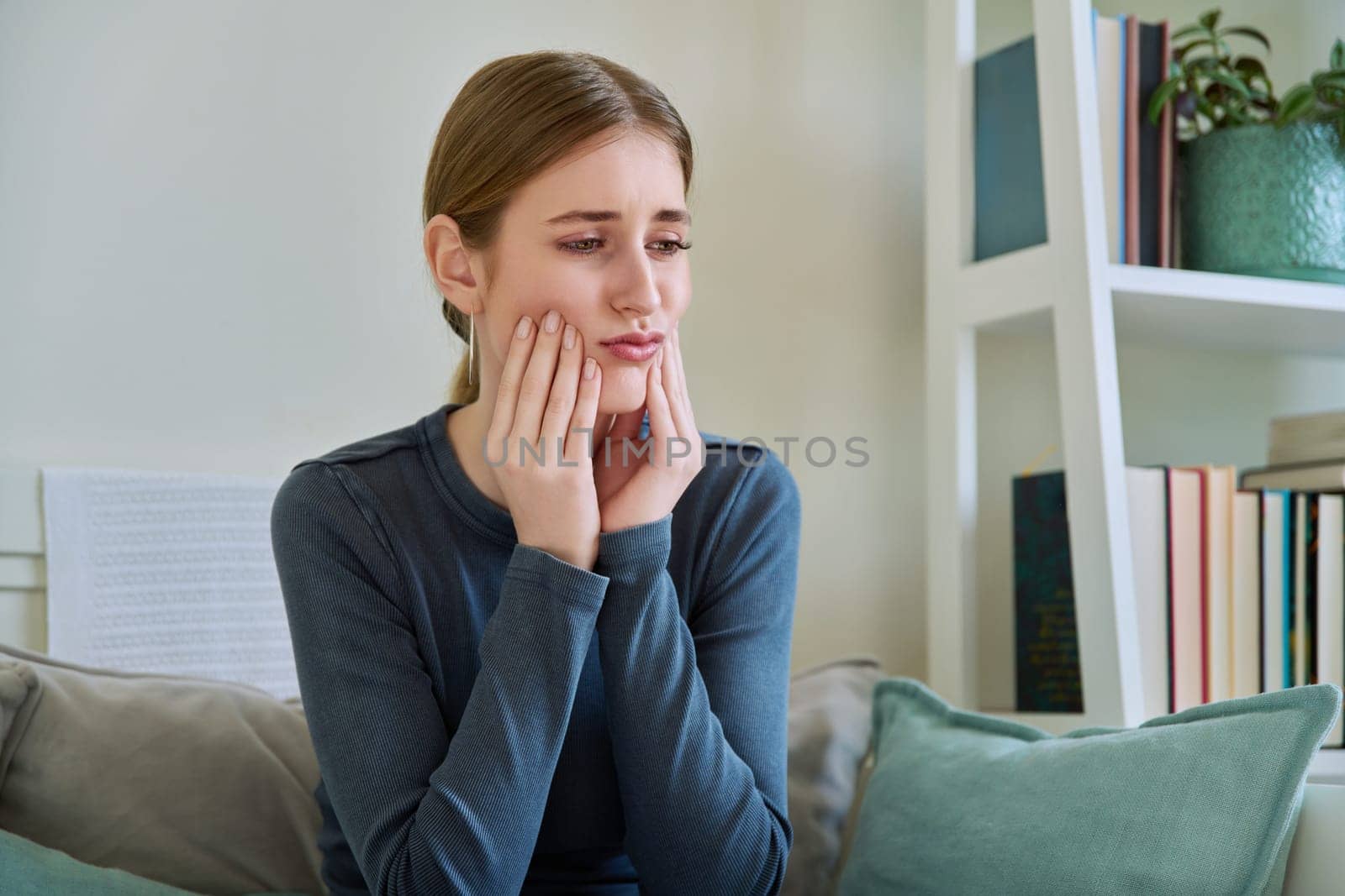 Young teenage female having toothache, suffering sad holding hands on face, sitting on couch at home. Teeth dentistry, dental, health, youth concept