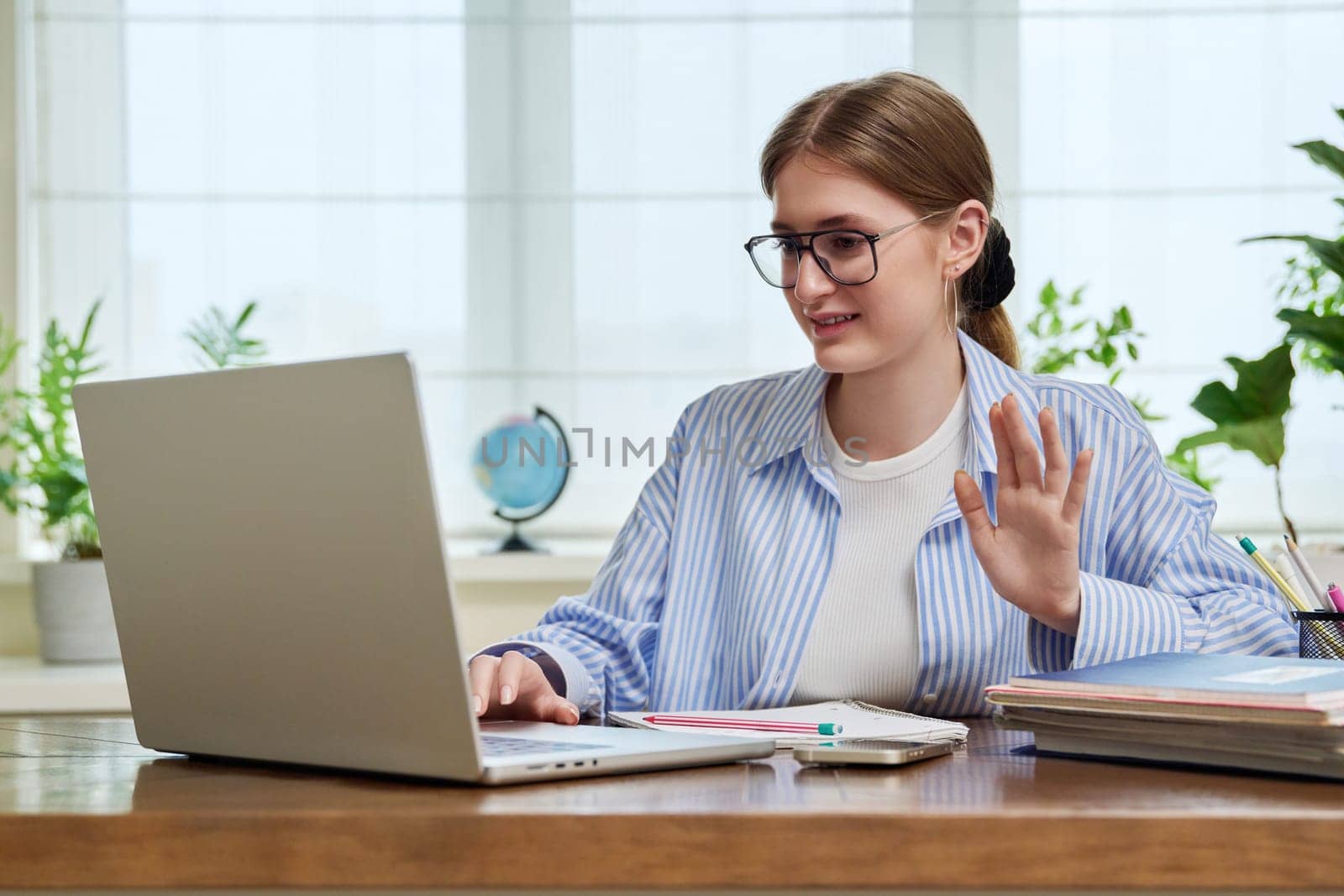 Young female student sitting at home at desk looking talking in web camera of computer laptop. Girl 17,18 years old studying remotely, video conference chat call, e-learning technology education