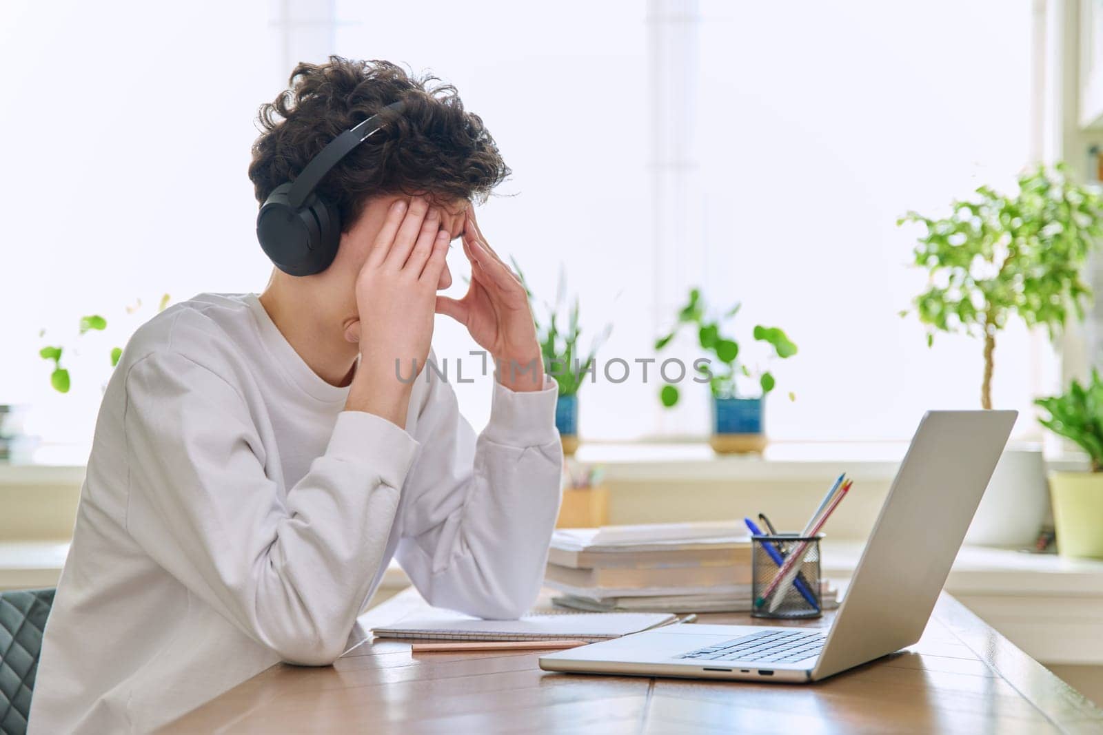 Sad upset young guy student in headphones sitting at home at desk with computer laptop, touching head with hands. Problems headaches troubles in college difficulties in study mental stress depression
