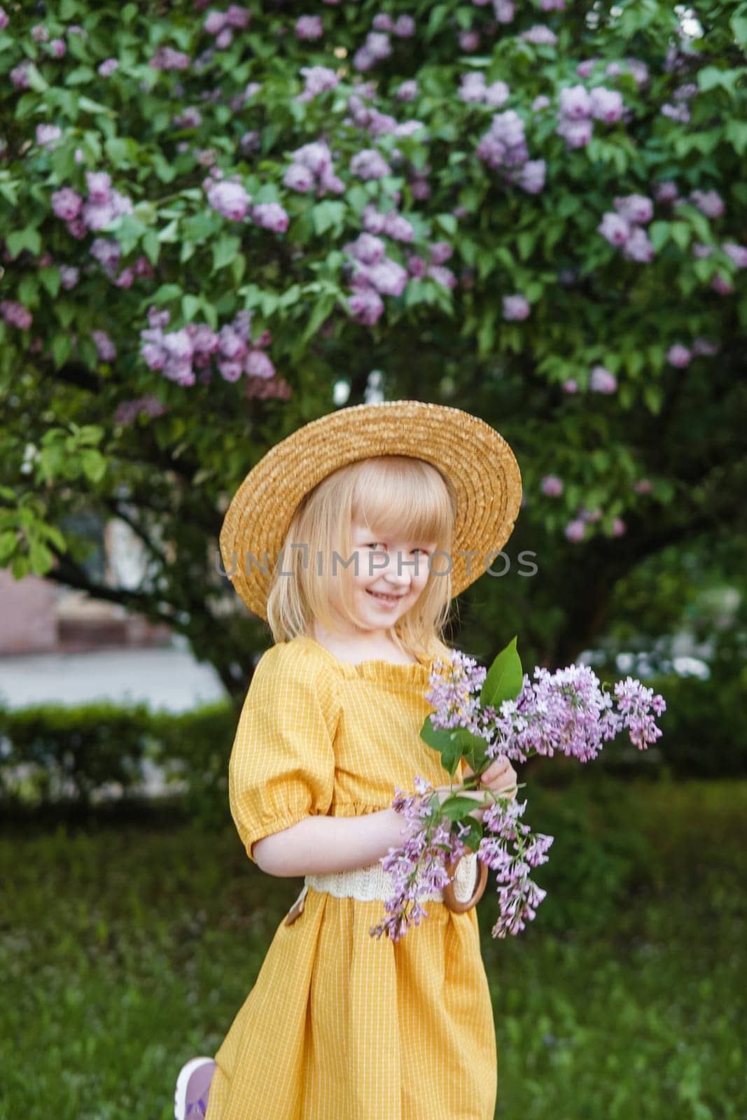 A little girl in a yellow dress and straw hat wearing a bouquet of lilacs. A walk in a spring park, blossoming lilacs