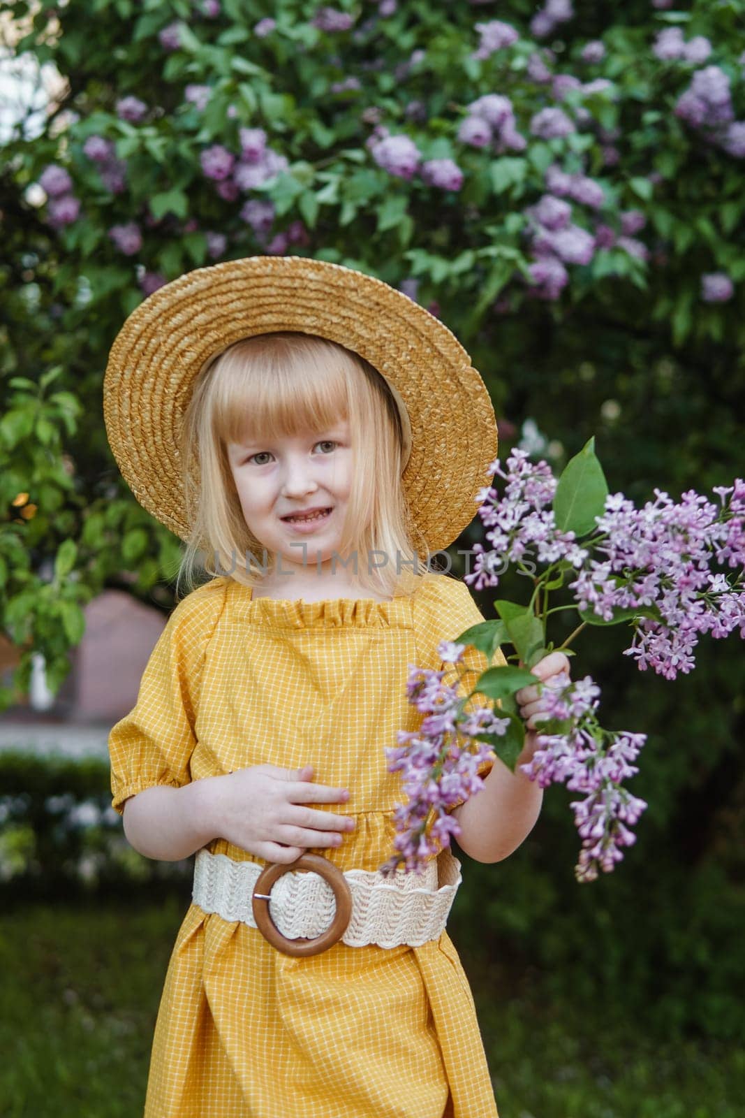 A little girl in a yellow dress and straw hat wearing a bouquet of lilacs. A walk in a spring park, blossoming lilacs