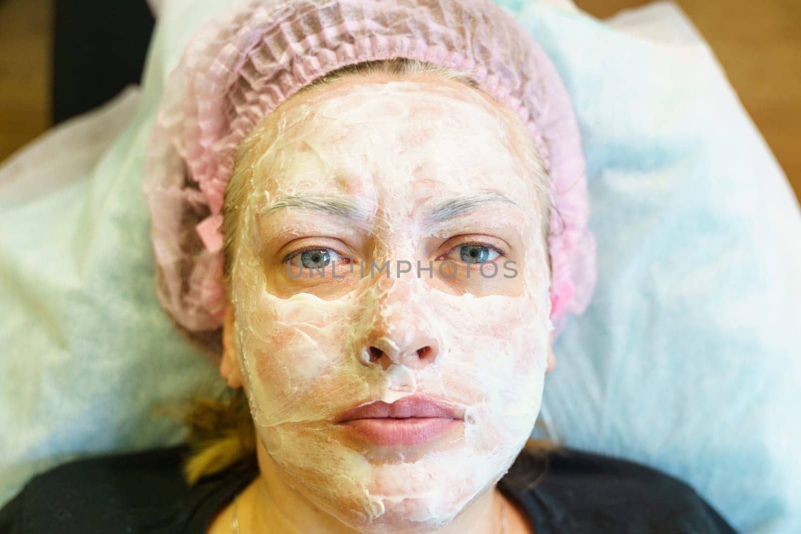 A woman is laying on a bed with a white facial mask applied, showing a skincare routine at home.