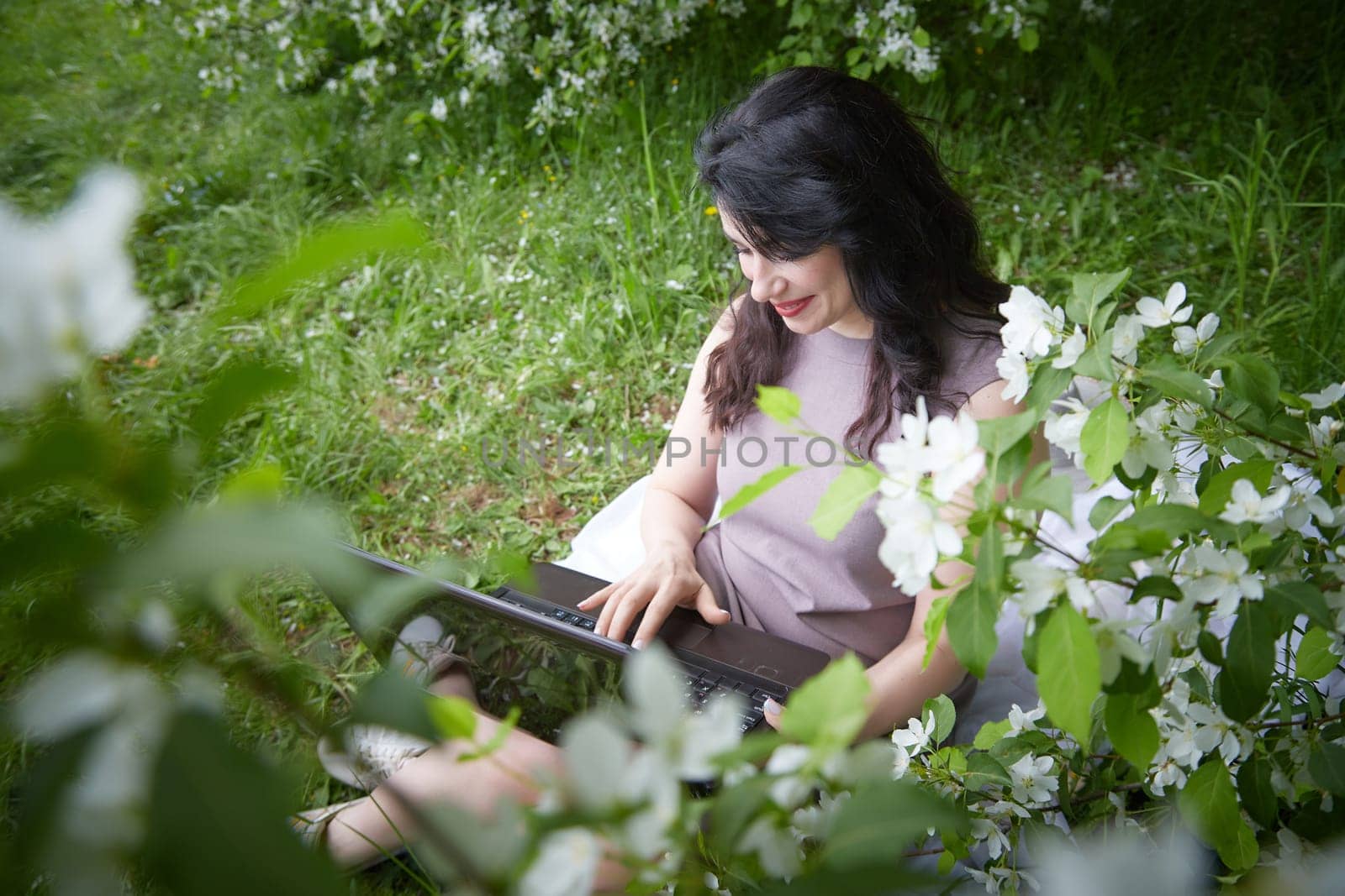 Girl engages with laptop amidst lush greenery. Freelancer Woman Working on Laptop Outdoors in a Blooming Garden