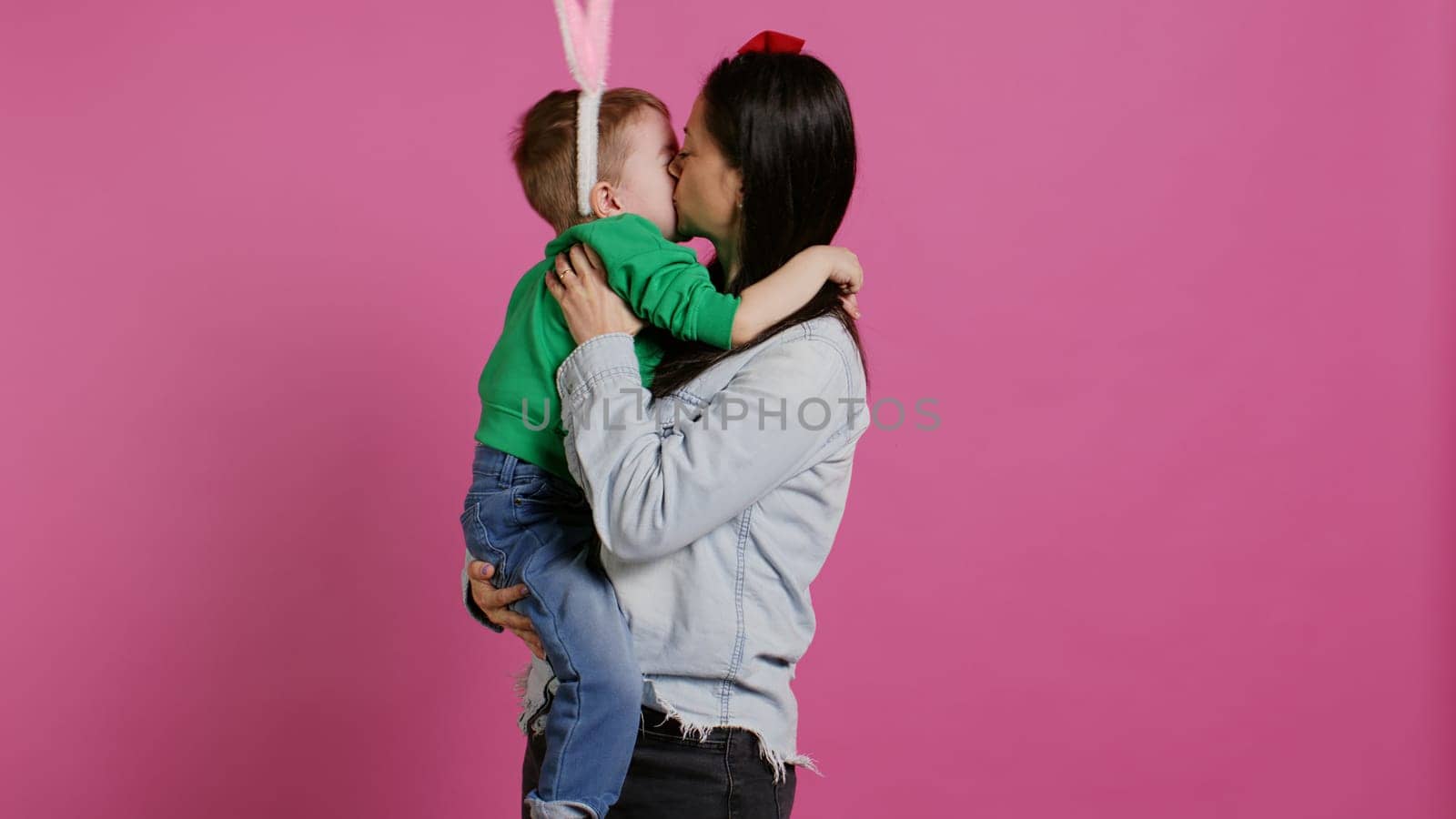 Cute mother and child showing love to each other in studio, mom hugging and kissing her small toddler son with bunny ears. Adorable little family laughing together and having fun. Camera B.