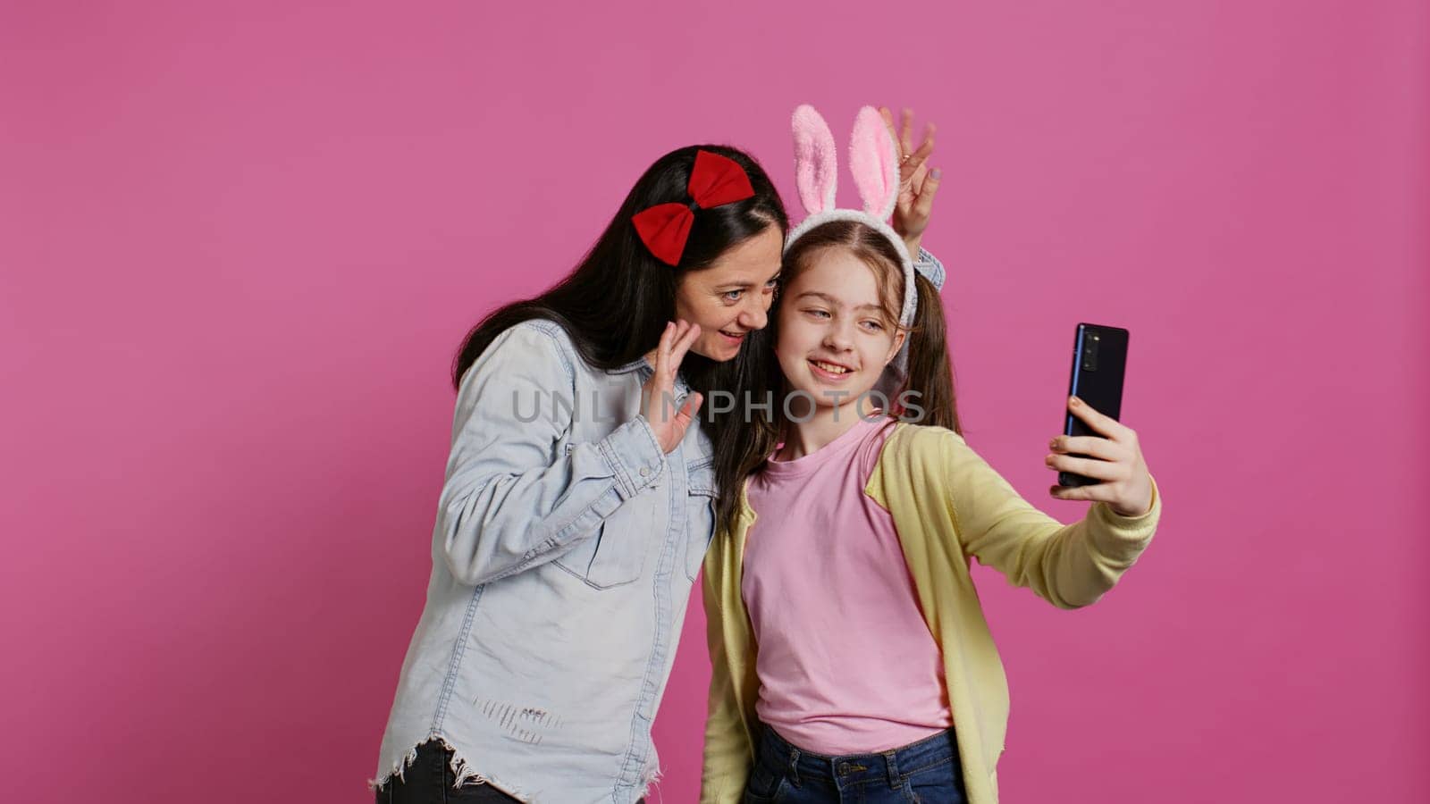 Playful cheery mother and girl smiling for photos on smartphone, having fun embracing each other in studio. Little child an her mom posing for pictures, fooling around. Camera B.