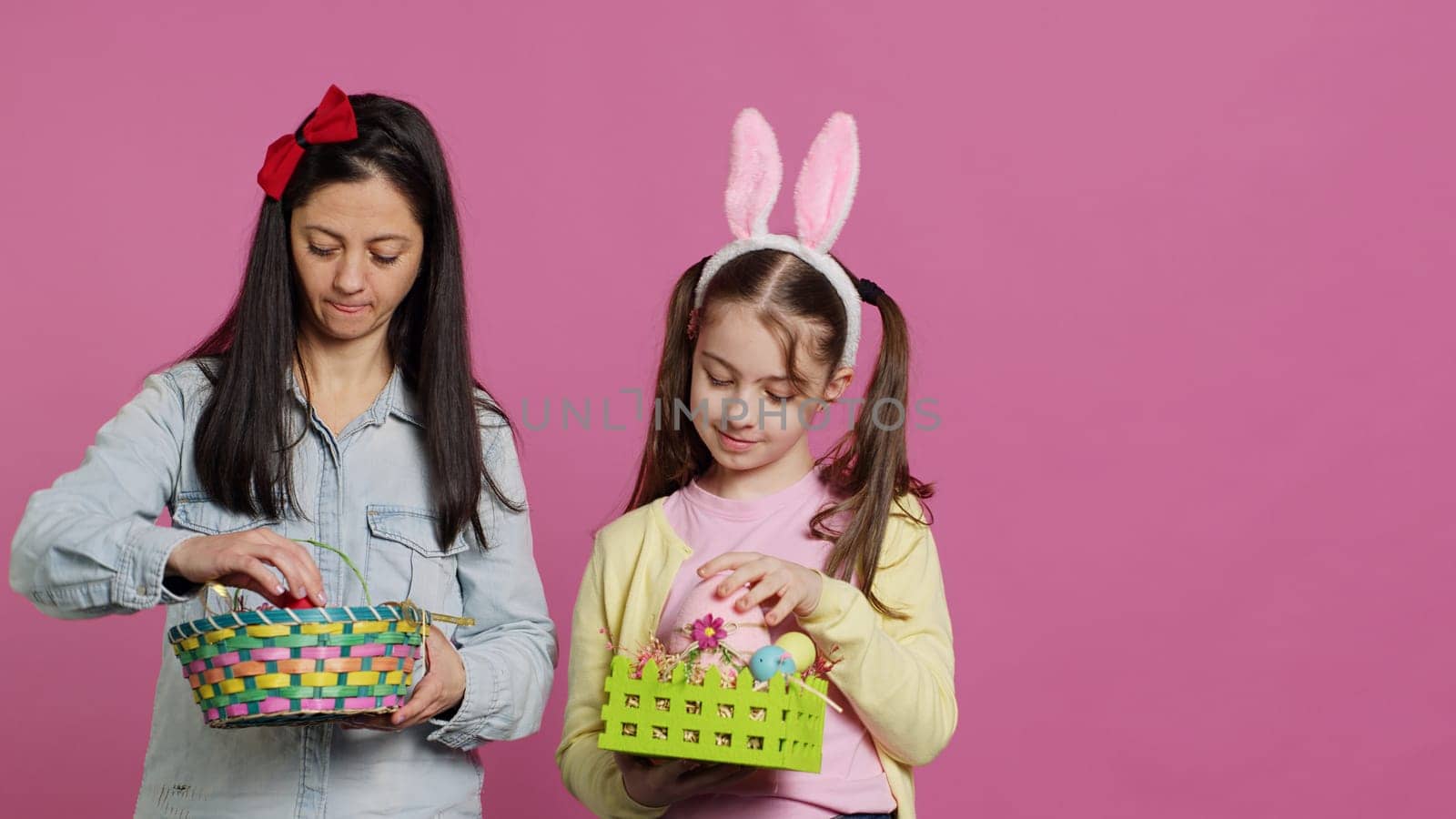 Joyful confident child and mother showing easter baskets on camera, decorating festive arrangements for spring holiday. Happy schoolgirl with bunny ears posing with her mom in studio. Camera A.