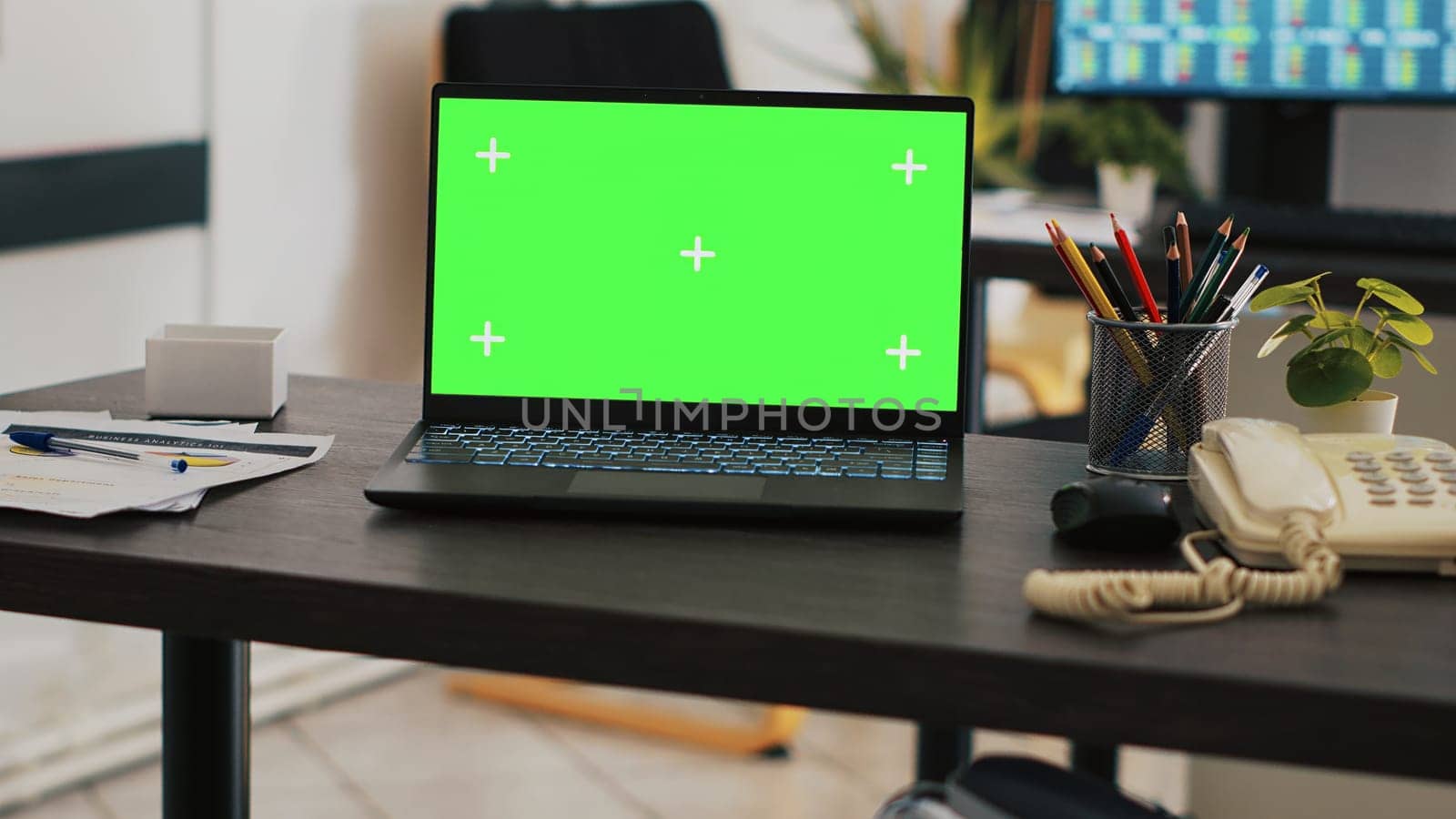 Focus on green screen laptop on office desk with computer in blurry background showing stock exchange data. Close up of mockup notebook and trading platform displayed on PC screen in the back