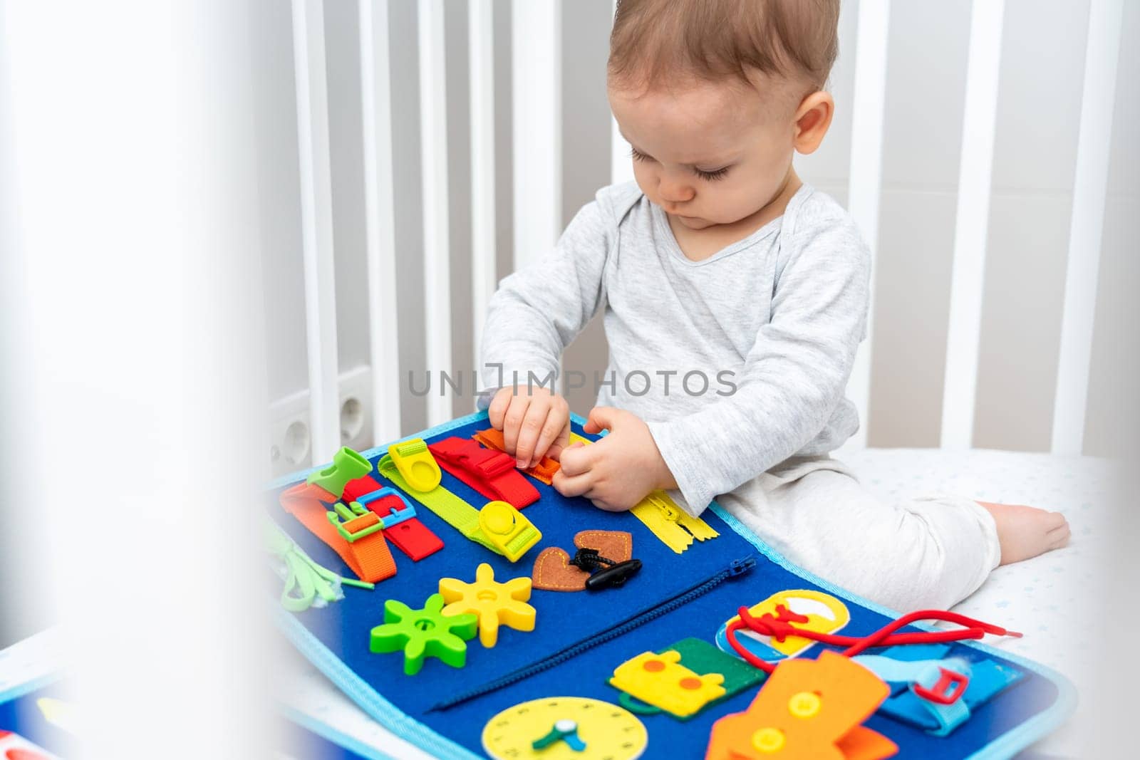Baby sitting in a crib playing with montessori busy book. Concept of keeping children from screen by activity books and quiet books by Mariakray
