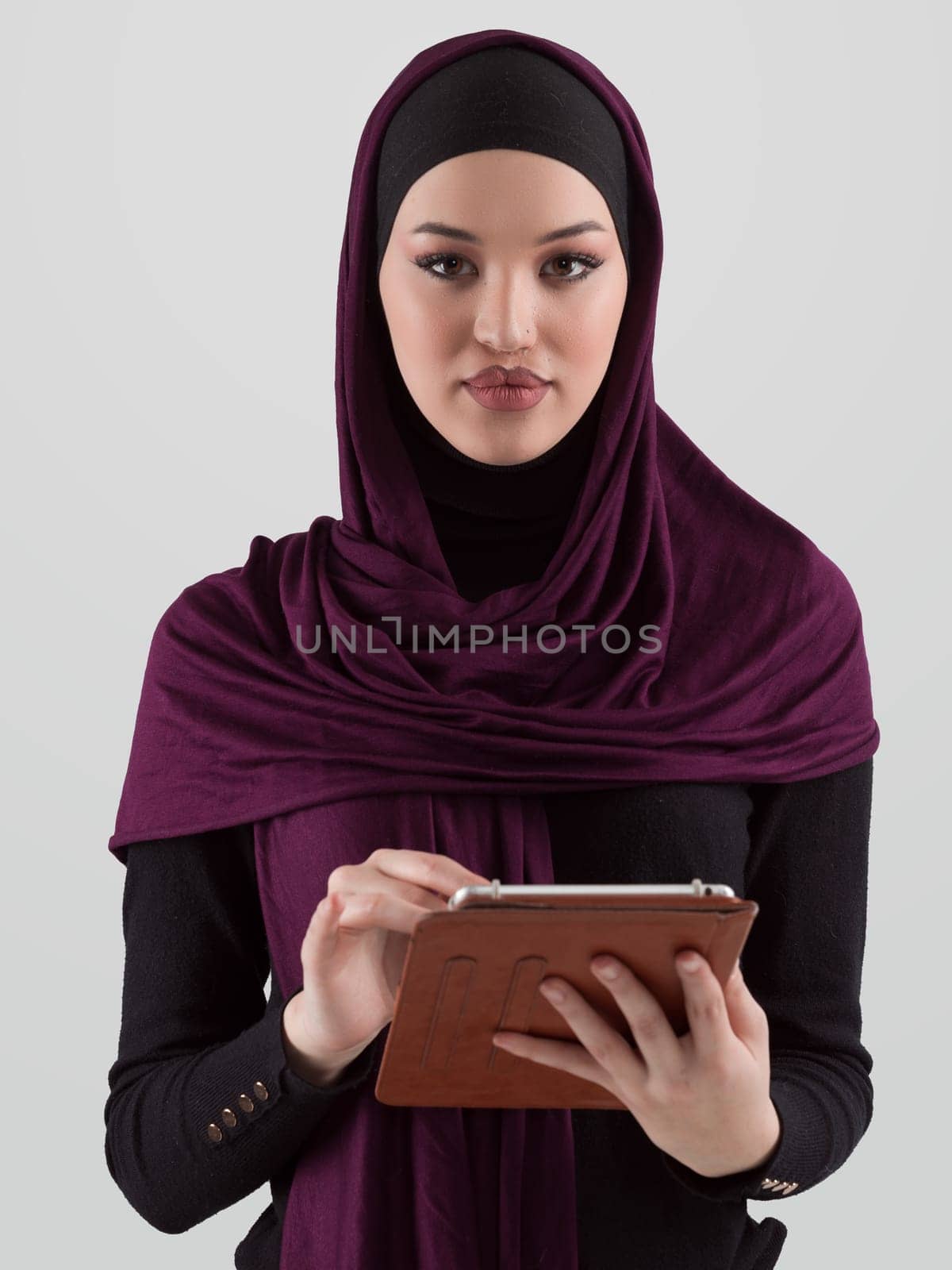 Beautiful and modern young Arabic businesswoman in hijab using a tablet computer while posing on light background and smiling at the camera. Business diversity concept, Muslim lady. by dotshock