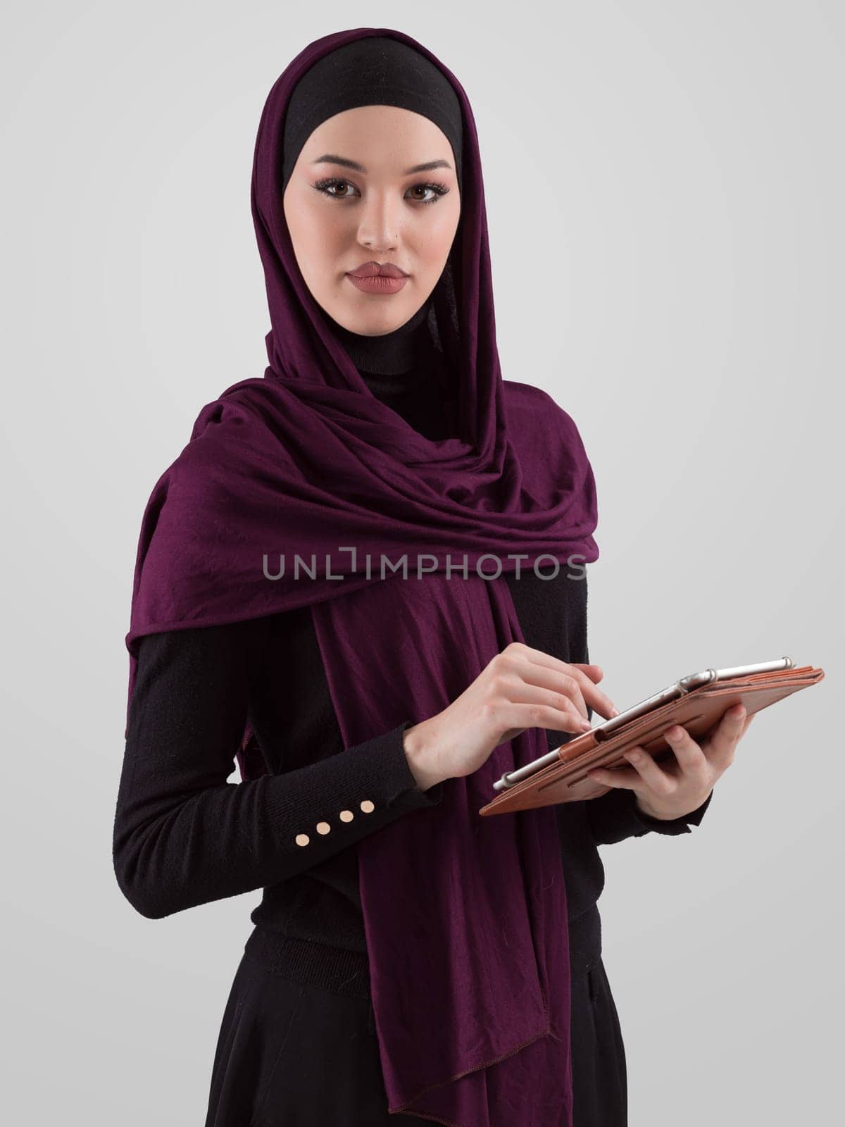 Beautiful and modern young Arabic businesswoman in hijab using a tablet computer while posing on light background and smiling at the camera. Business diversity concept, Muslim lady. by dotshock