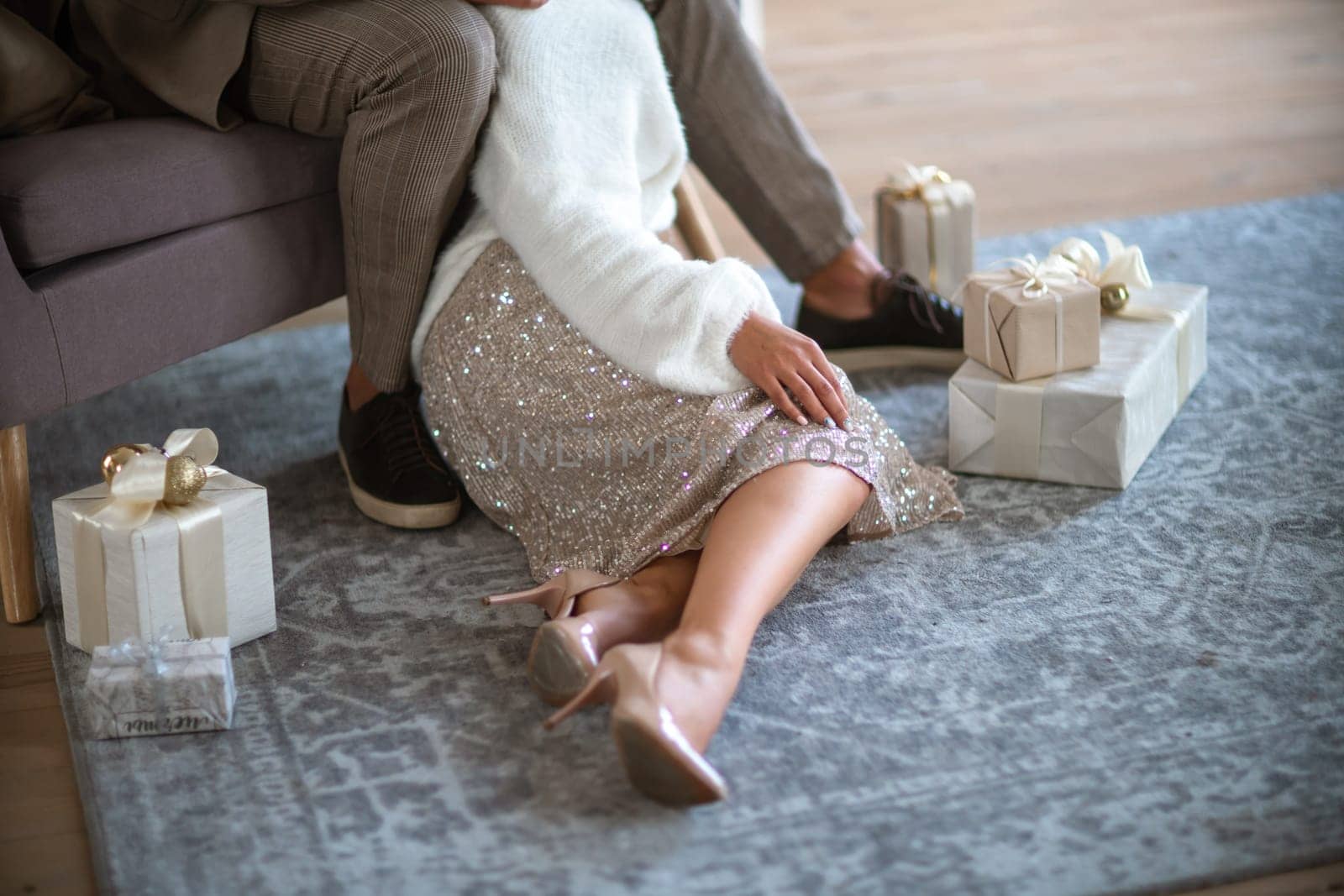 A romantic couple, a man sitting on the sofa and a woman on the carpet next to surrounded by gifts on a gray carpet. She is wearing a light skirt and beige high-heeled shoes