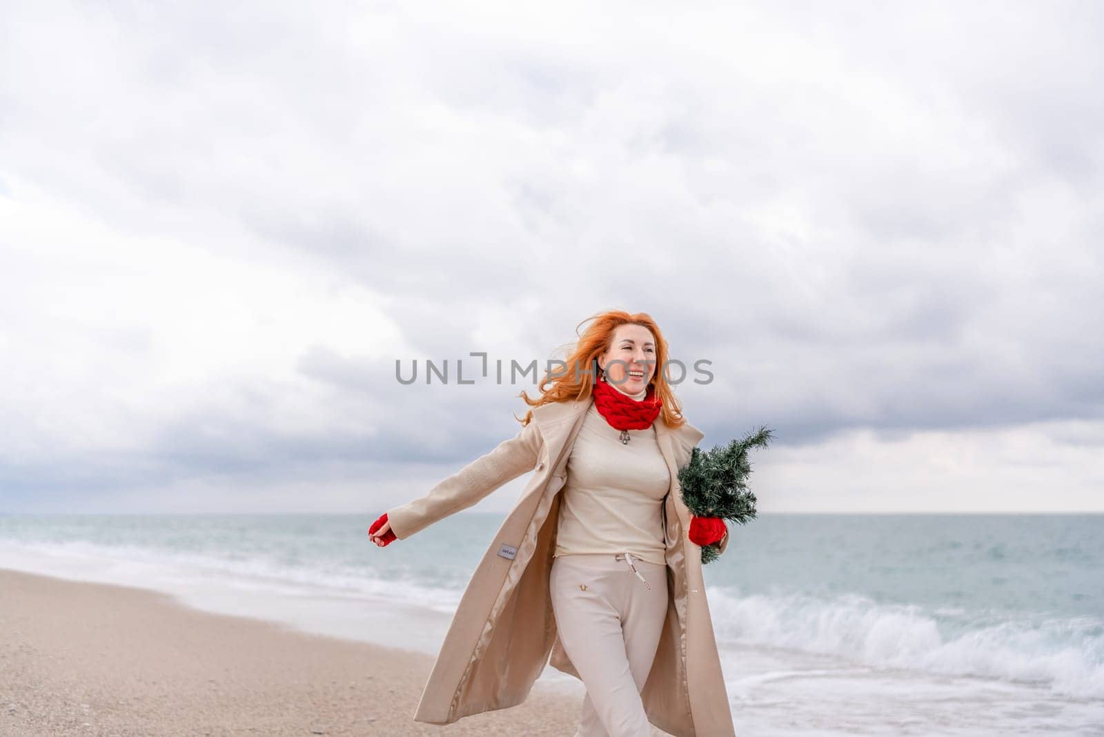 Redhead woman Christmas tree sea. Christmas portrait of a happy redhead woman walking along the beach and holding a Christmas tree in her hands. Dressed in a light coat, white suit and red mittens