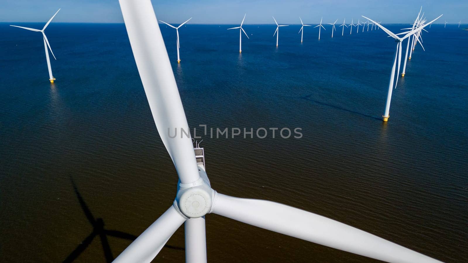 A group of windmill turbines on the ocean waters, their blades spinning in the gentle spring breeze of Flevoland, Netherlands. drone aerial view of windmill turbines green energy in the ocean