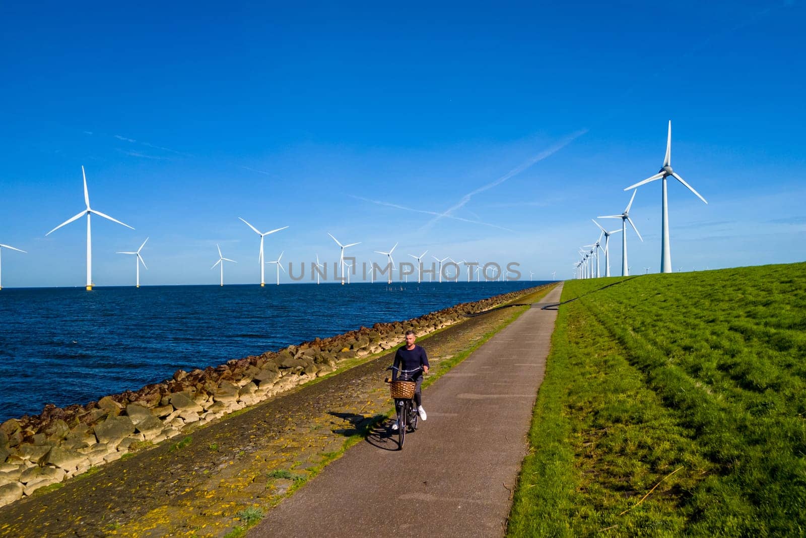 A man energetically cycling down a path lined with wind turbines in the Netherlands Flevoland during the vibrant spring season. men on electric bike with windmill turbines on the background