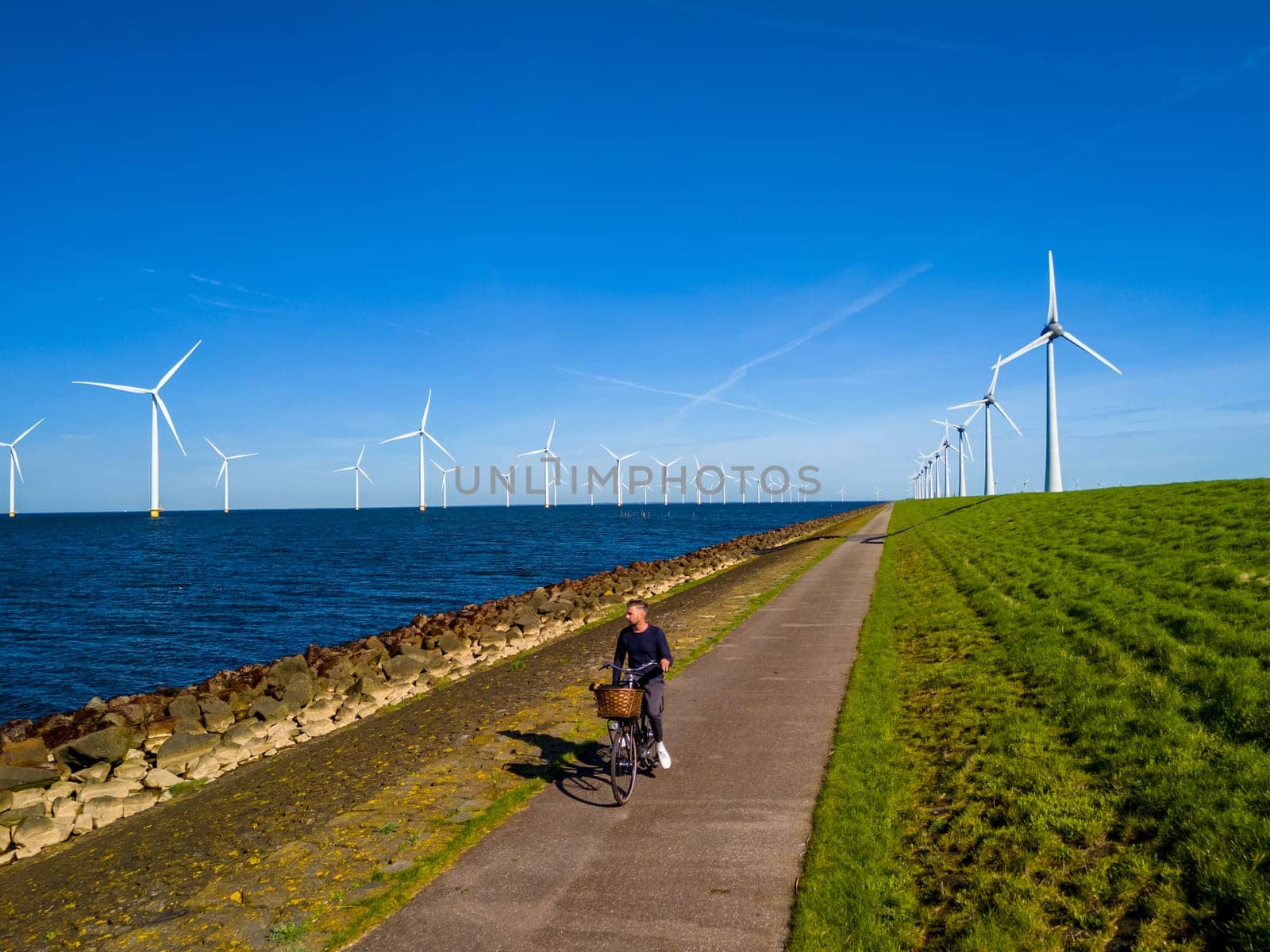 A man gracefully rides a bike down a picturesque path next to windmill turbines in Spring by fokkebok