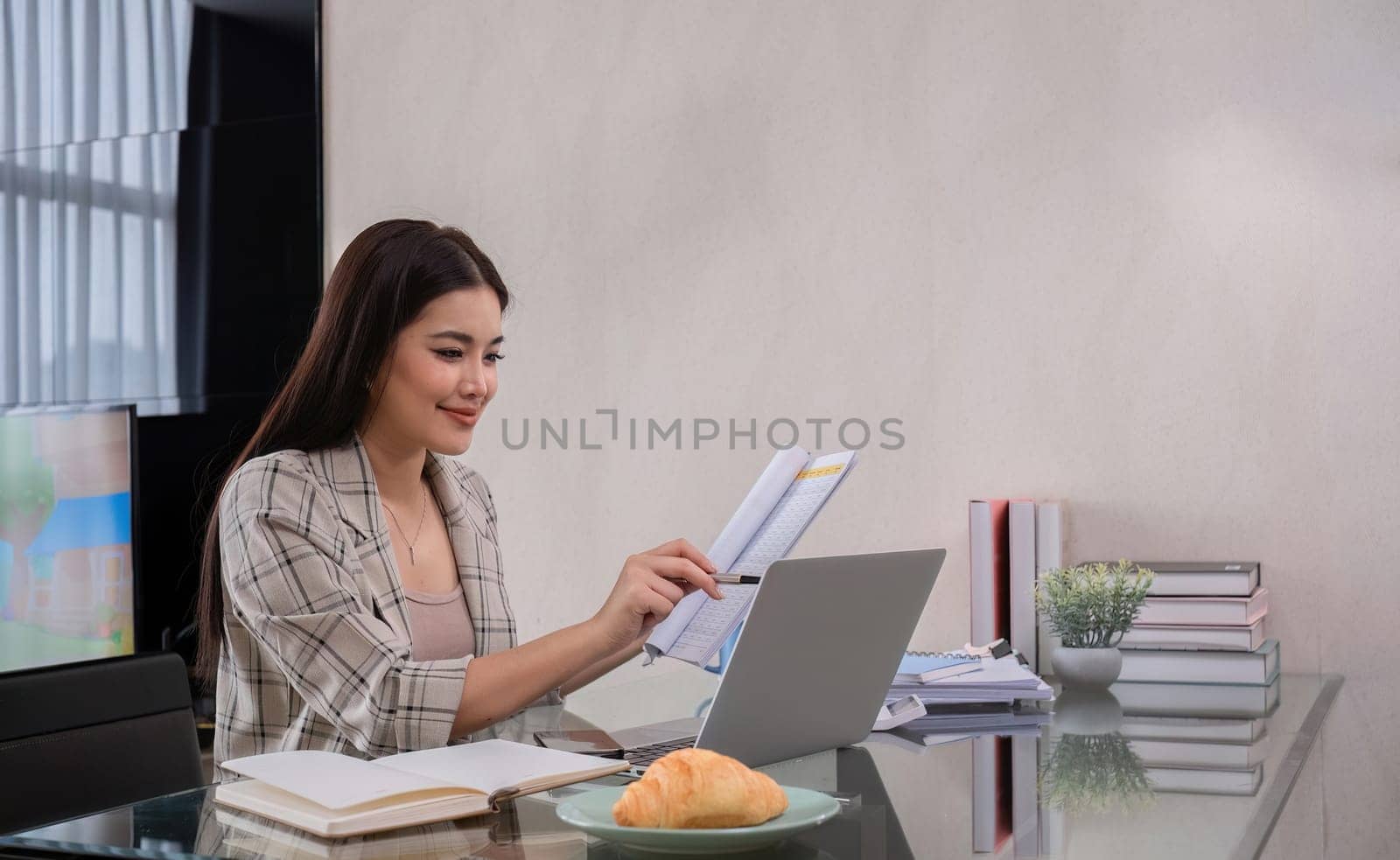 A middle-aged woman works from home at the dining room table with a laptop and a stack of documents next to her..