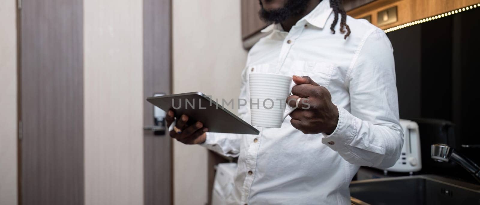 African American man working online on digital tablet and drinking coffee while at kitchen by nateemee