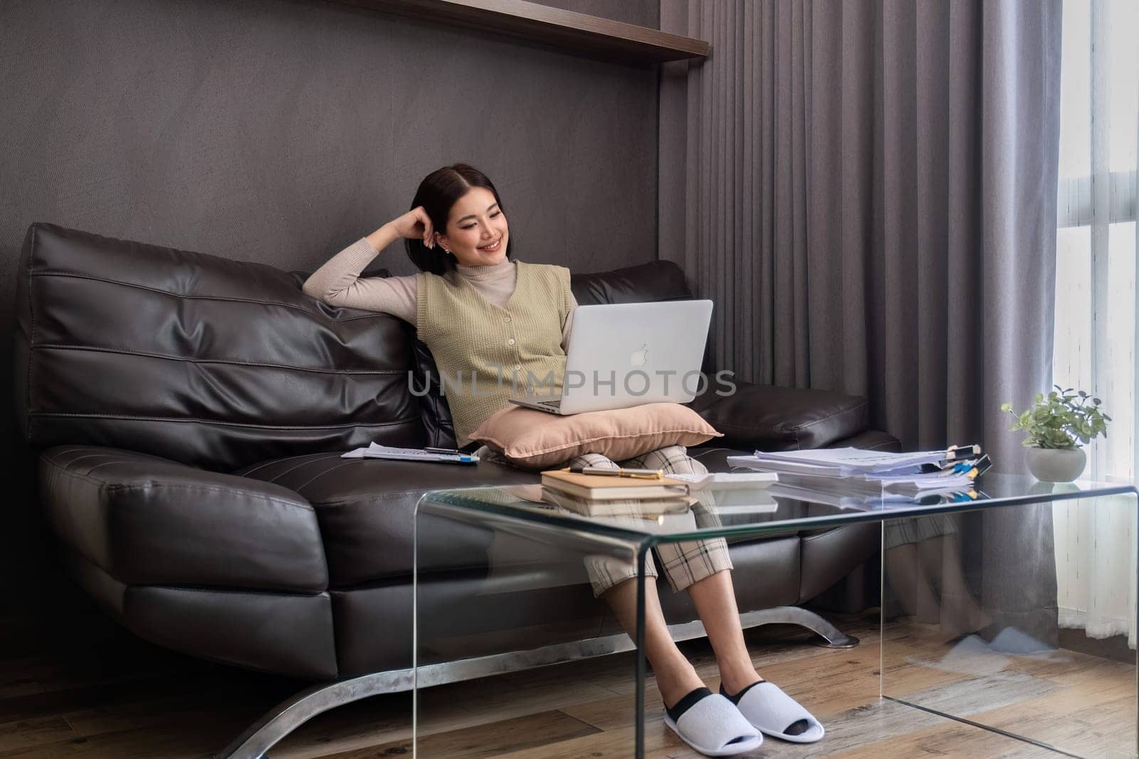 A middle-aged woman works from home at the dining room table with a laptop and a stack of documents next to her..