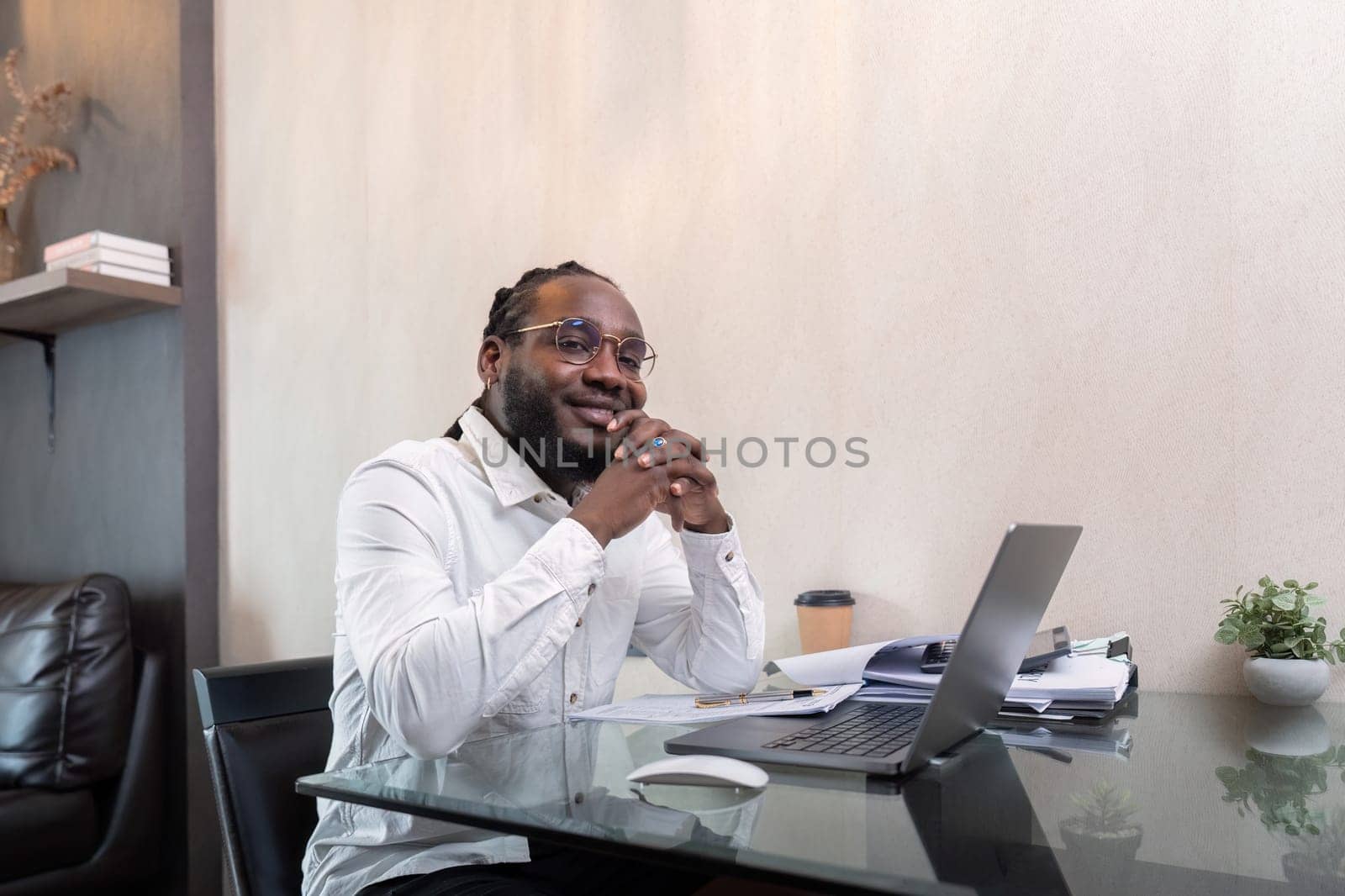 Smiling African American man sit at desk in home working on laptop, happy African American male look at camera posing, busy using modern computer gadget at home.