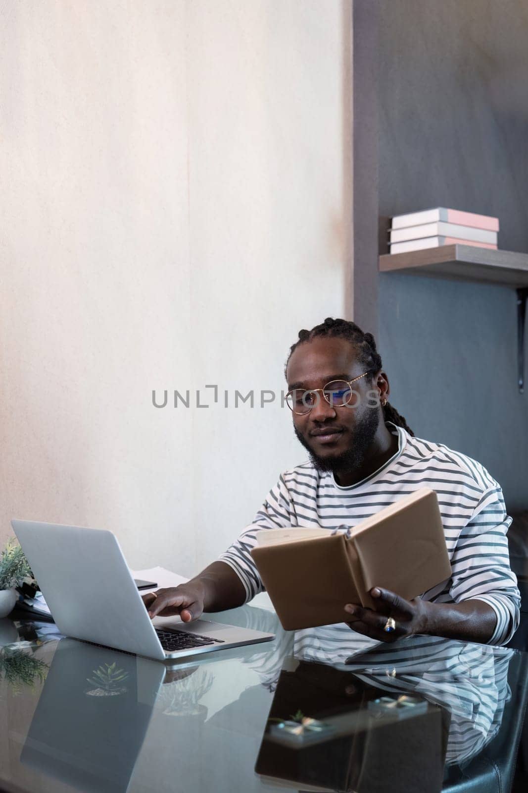 Young african man using laptop at home, black male looking at read book relaxing on leisure with work sit on glass table in living room.