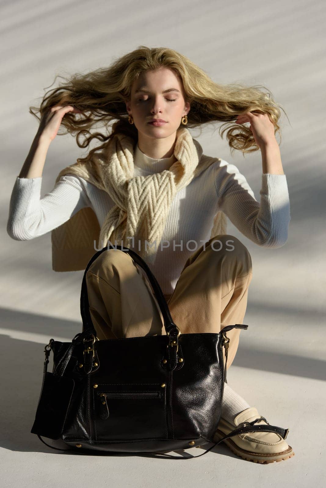 beautiful curly blond hair woman posing with a small shopper bag sitting on the floor. Model wearing stylish white sweater, classic trousers and loafer shoes