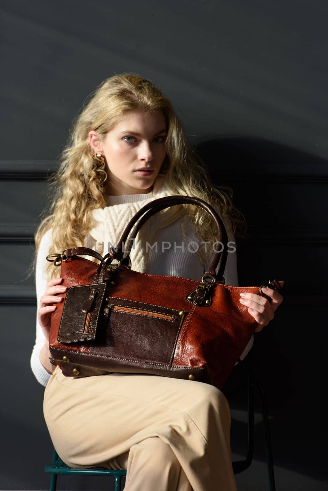 beautiful curly blond hair woman posing with a brown shopper bag near gray wall. Model wearing stylish white sweater and classic trousers.
