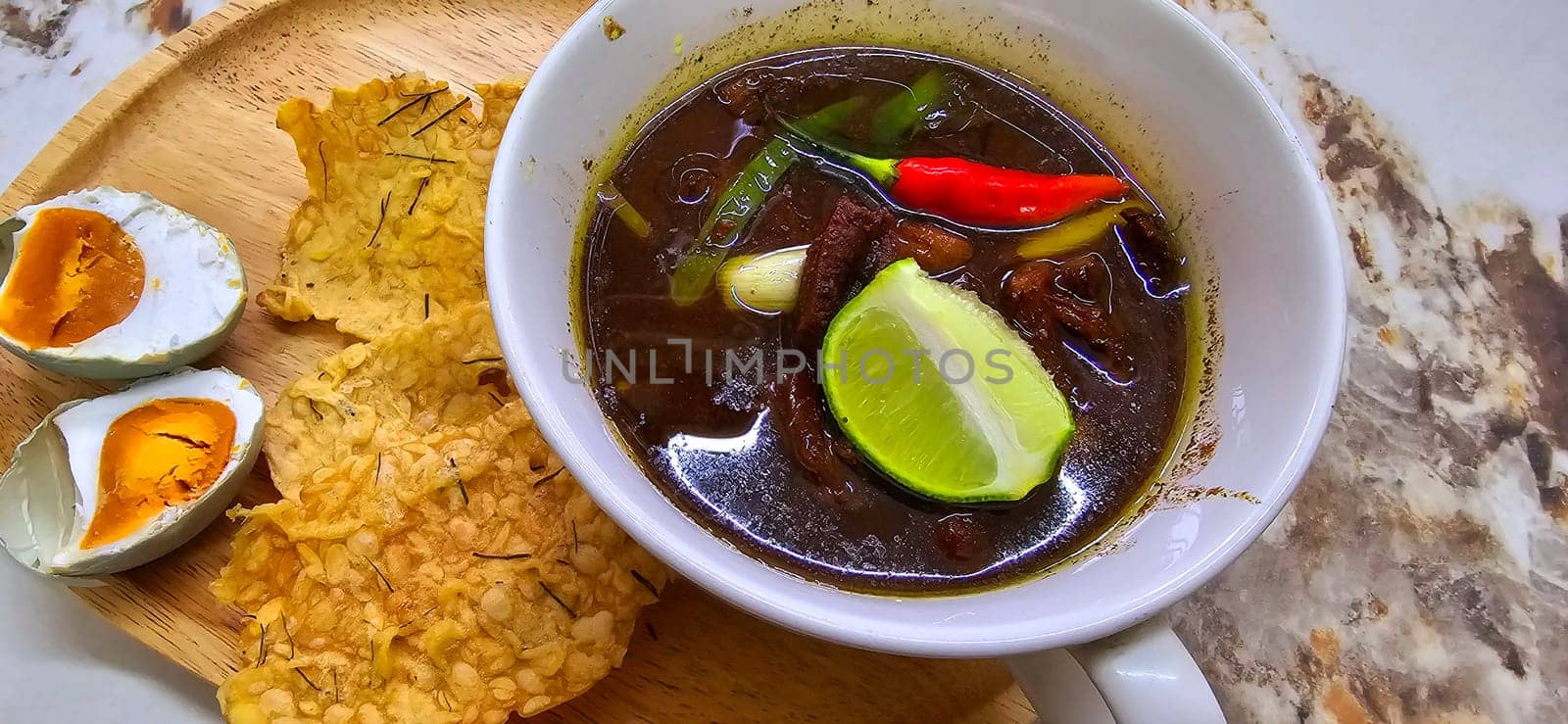 Rawon or Indonesian black beef soup, black color are from indonesian nut called kluwek. Served with lime, chili paste, salted egg, and tempe cracker. Perfect for recipe, article, or any cooking contents or blog
