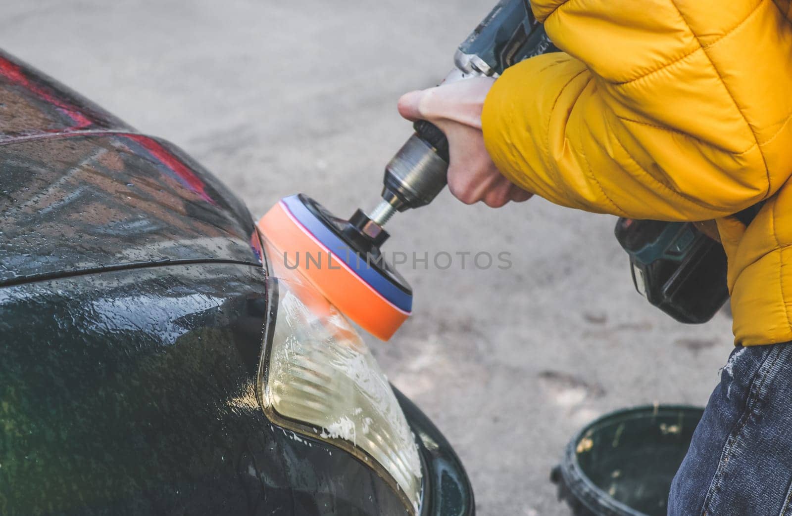 The hands of a young caucasian guy are drilling a drill with a disk with a sponge and polishing the headlight of his car with a detergent soap, close-up side view.The concept of a home car wash and cleaning with polishing car headlights.