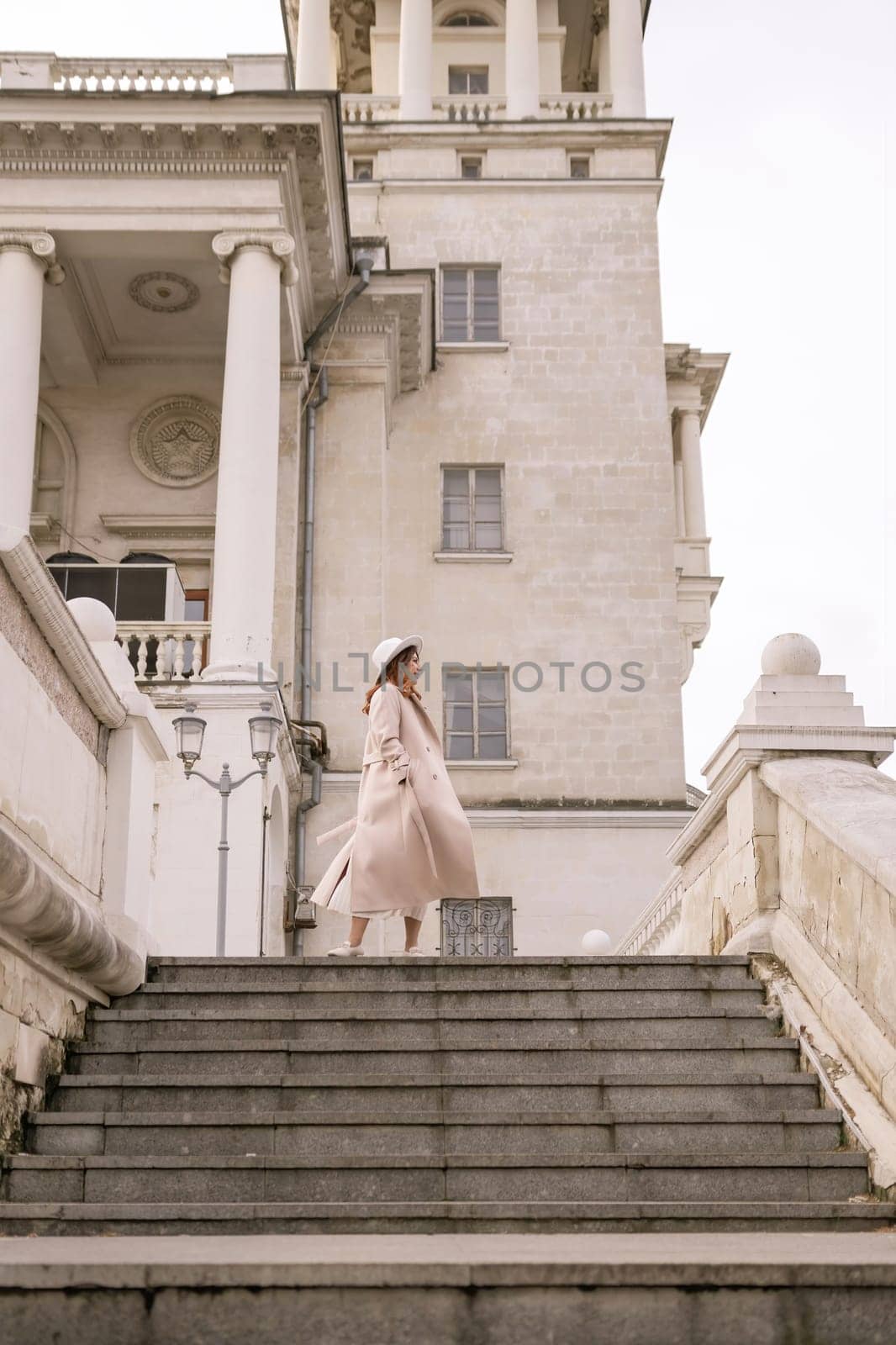 woman in elegant coat and hat against an intricate architectural backdrop, harmoniously blending modern fashion with historical allure. The soft daylight adds to its timeless appeal. by Matiunina