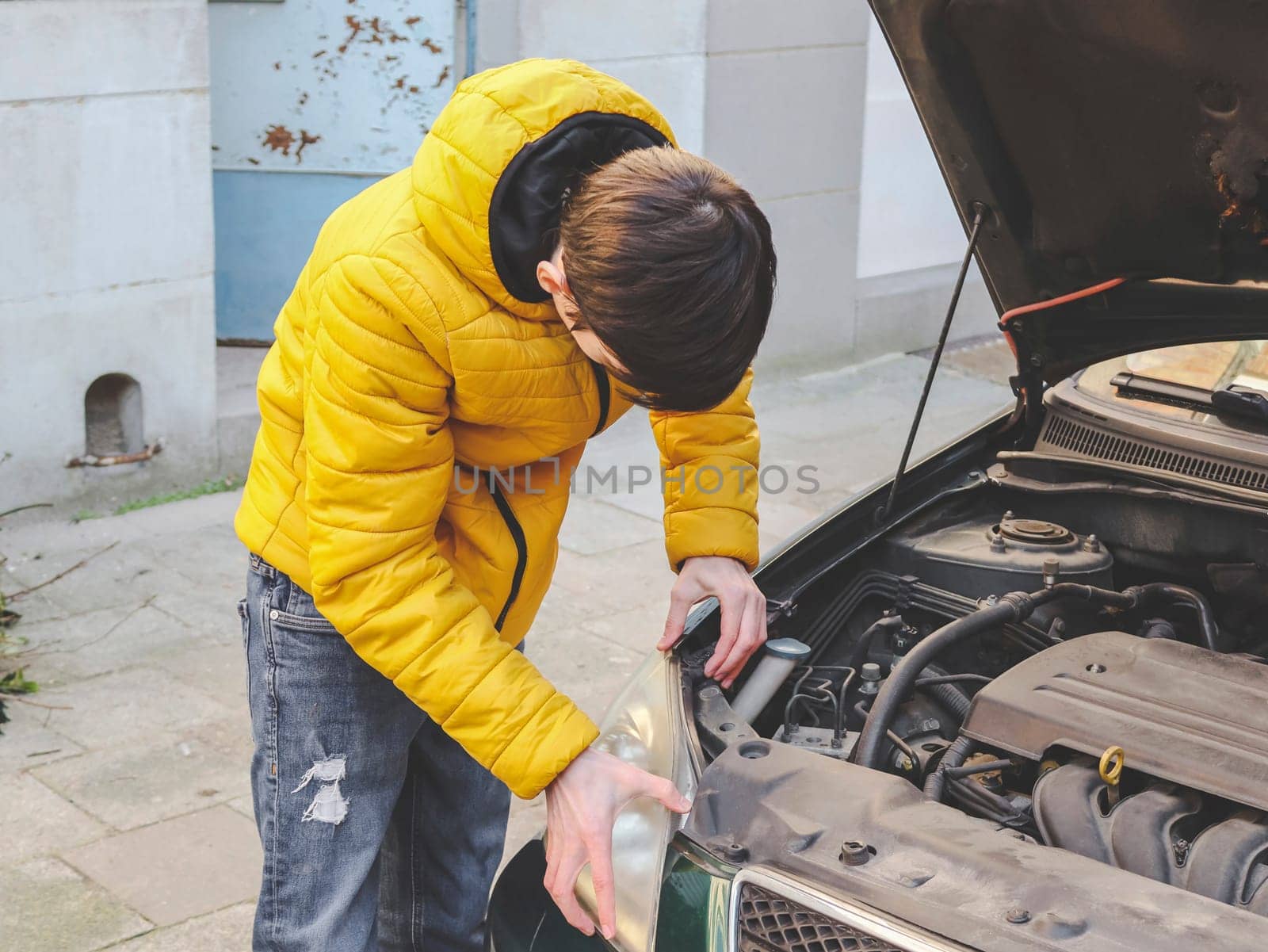A young caucasian guy in a yellow jacket inspects an internal breakdown with an open car hood of his car on a city street in front of an old house, close-up side view. Engine concept for home repair.