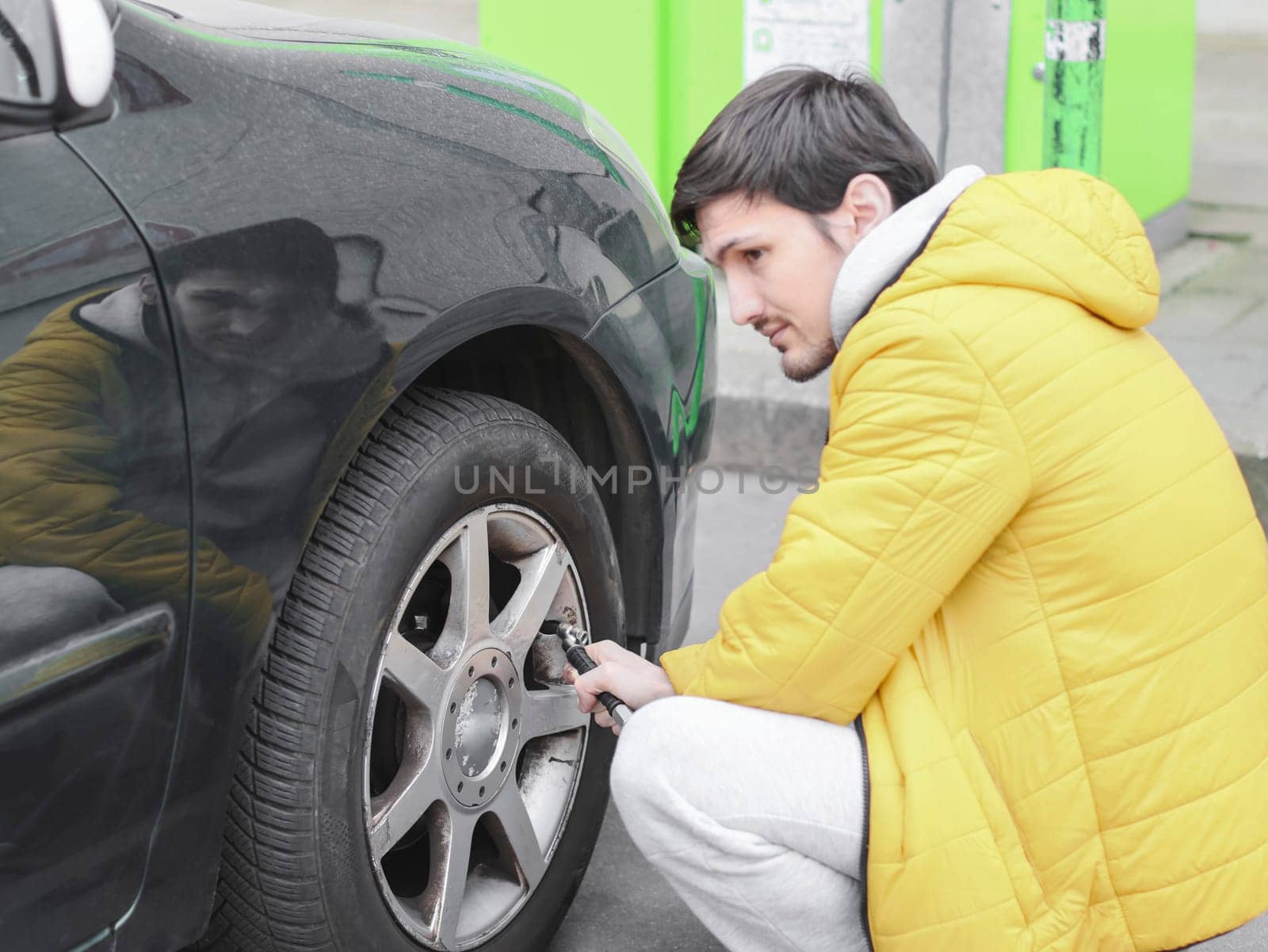 Young caucasian man in a yellow jacket and gray sweatpants, squatting thoughtfully, pumps air into the wheel of a car with an air compressor in a parking lot, side view with selective focus and closeup.Gas station concept.