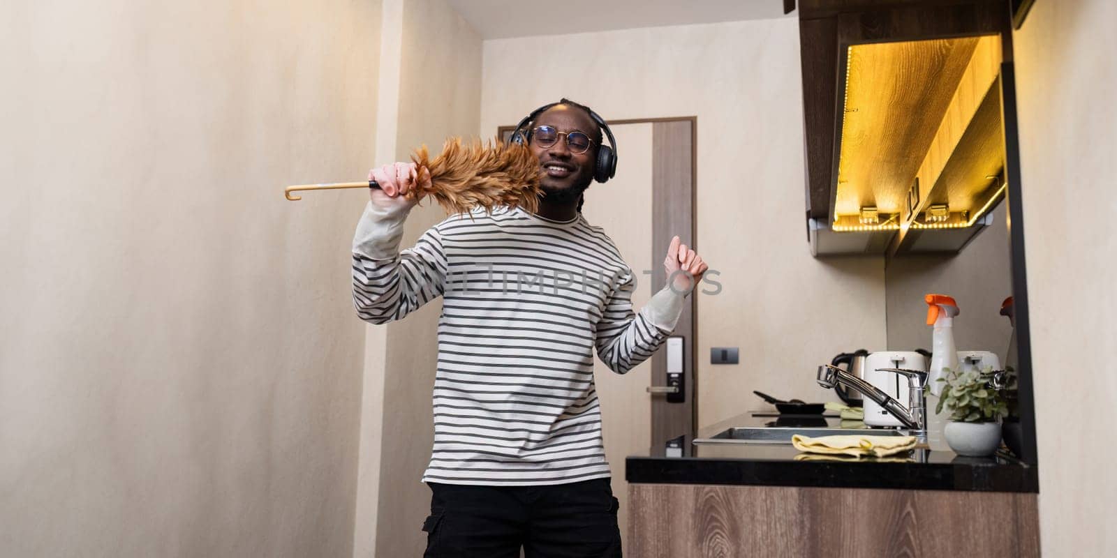 housework concept. African American joyful happy young guy cleaning the living room at home doing housework dancing having fun by itchaznong