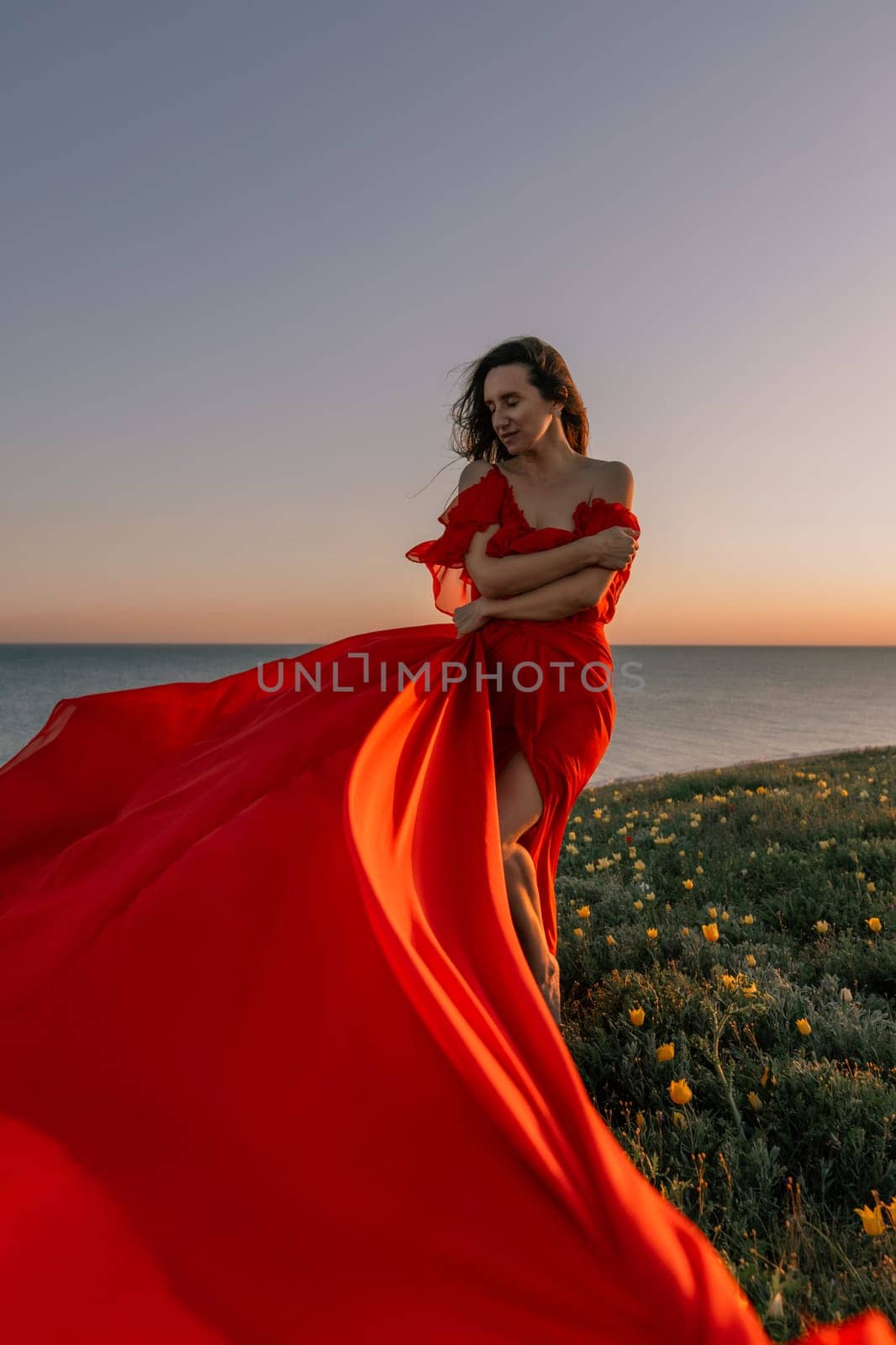 woman red dress standing grassy hillside. The sun is setting in the background, casting a warm glow over the scene. The woman is enjoying the beautiful view and the peaceful atmosphere