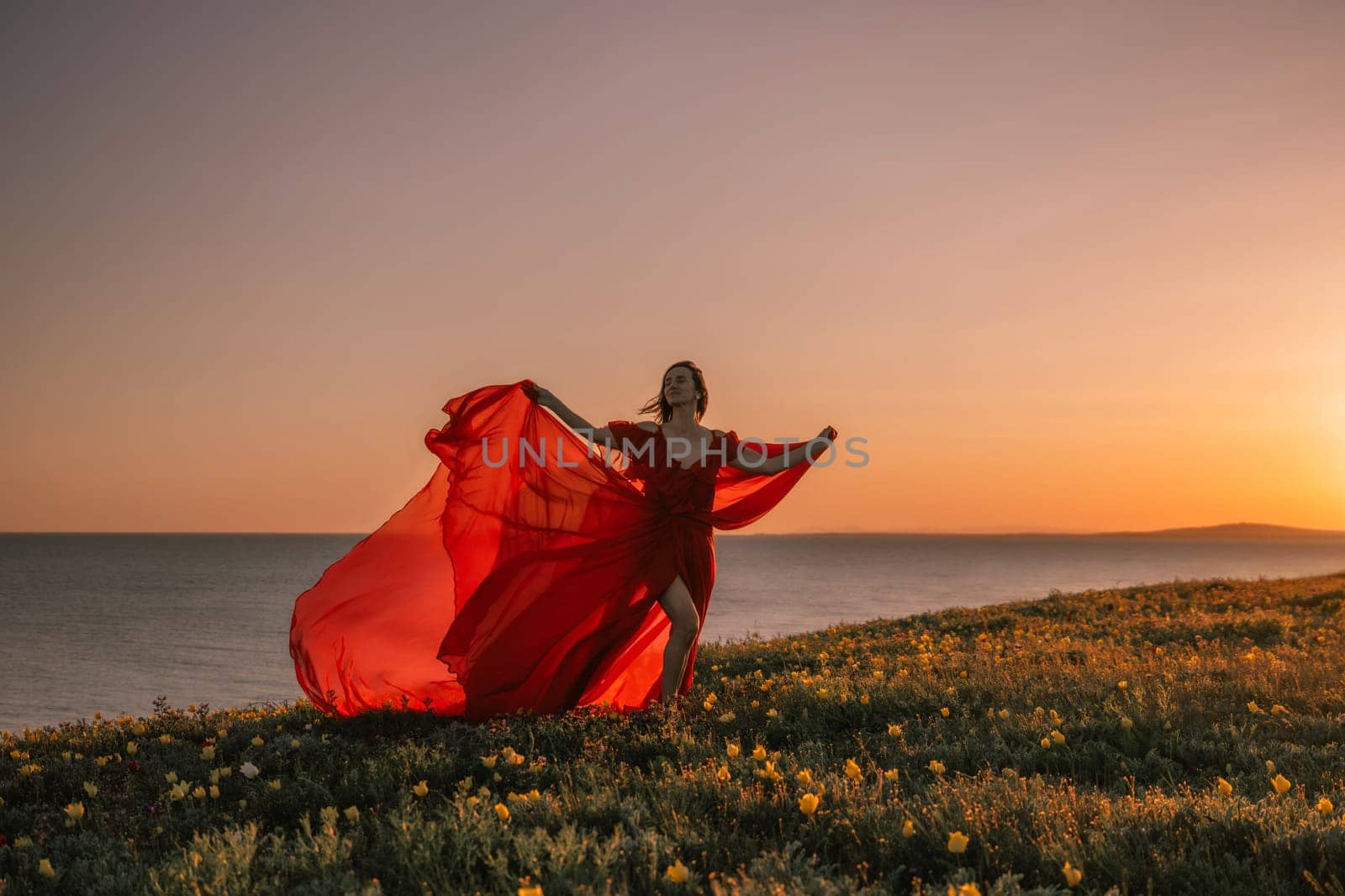 woman red dress is standing on a grassy hill overlooking the ocean. The sky is a beautiful mix of orange and pink hues, creating a serene and romantic atmosphere