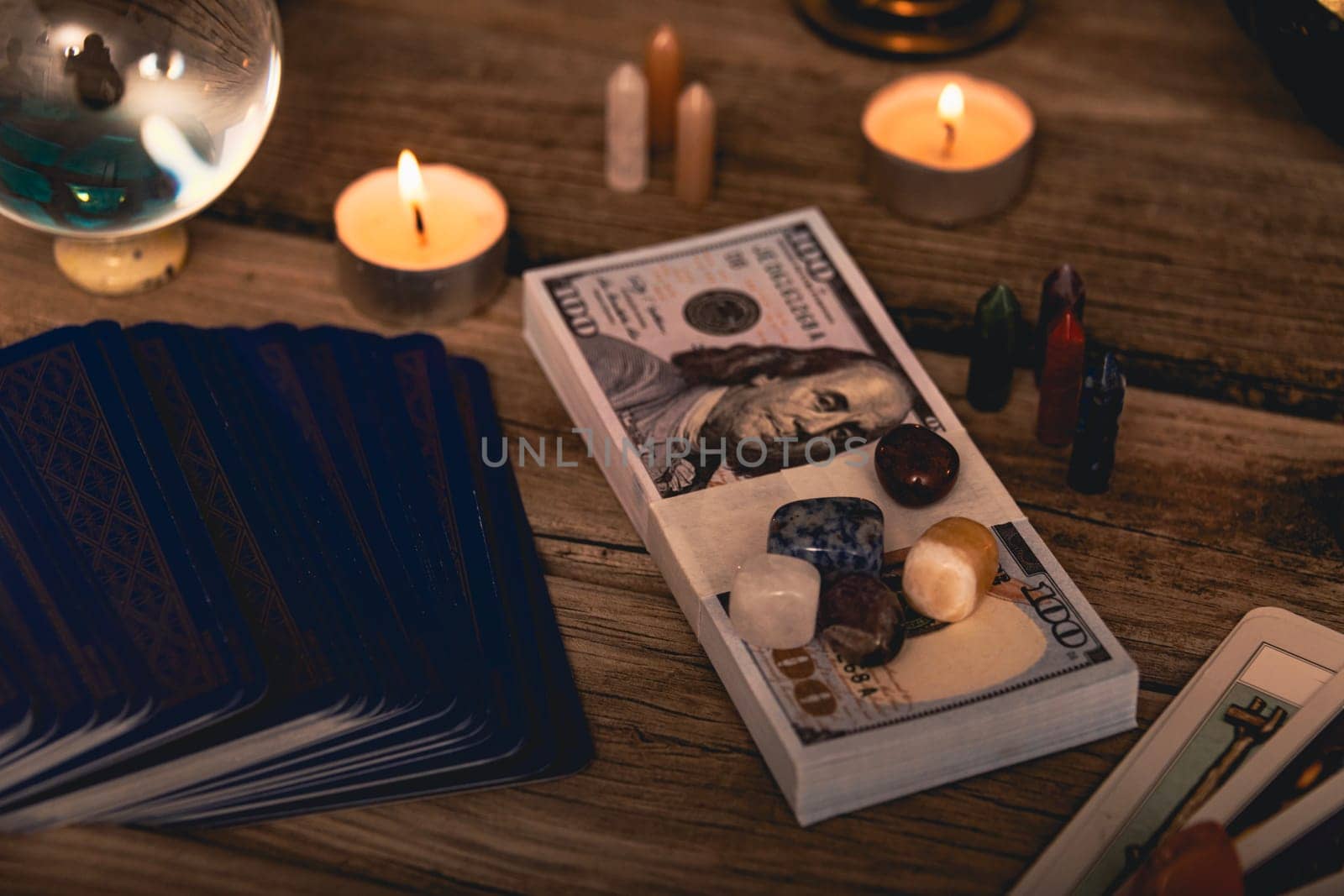 A tarot spread with The Fool card, hundred-dollar bills, and various crystals on a rustic wooden background with candles
